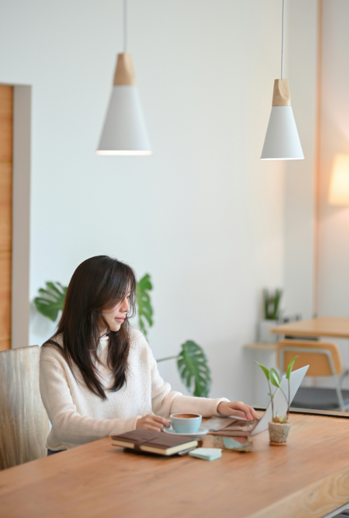 A woman sitting at a wooden table working on a laptop, with a cup, books, and a small potted plant in a bright, modern room with hanging pendant lights.
