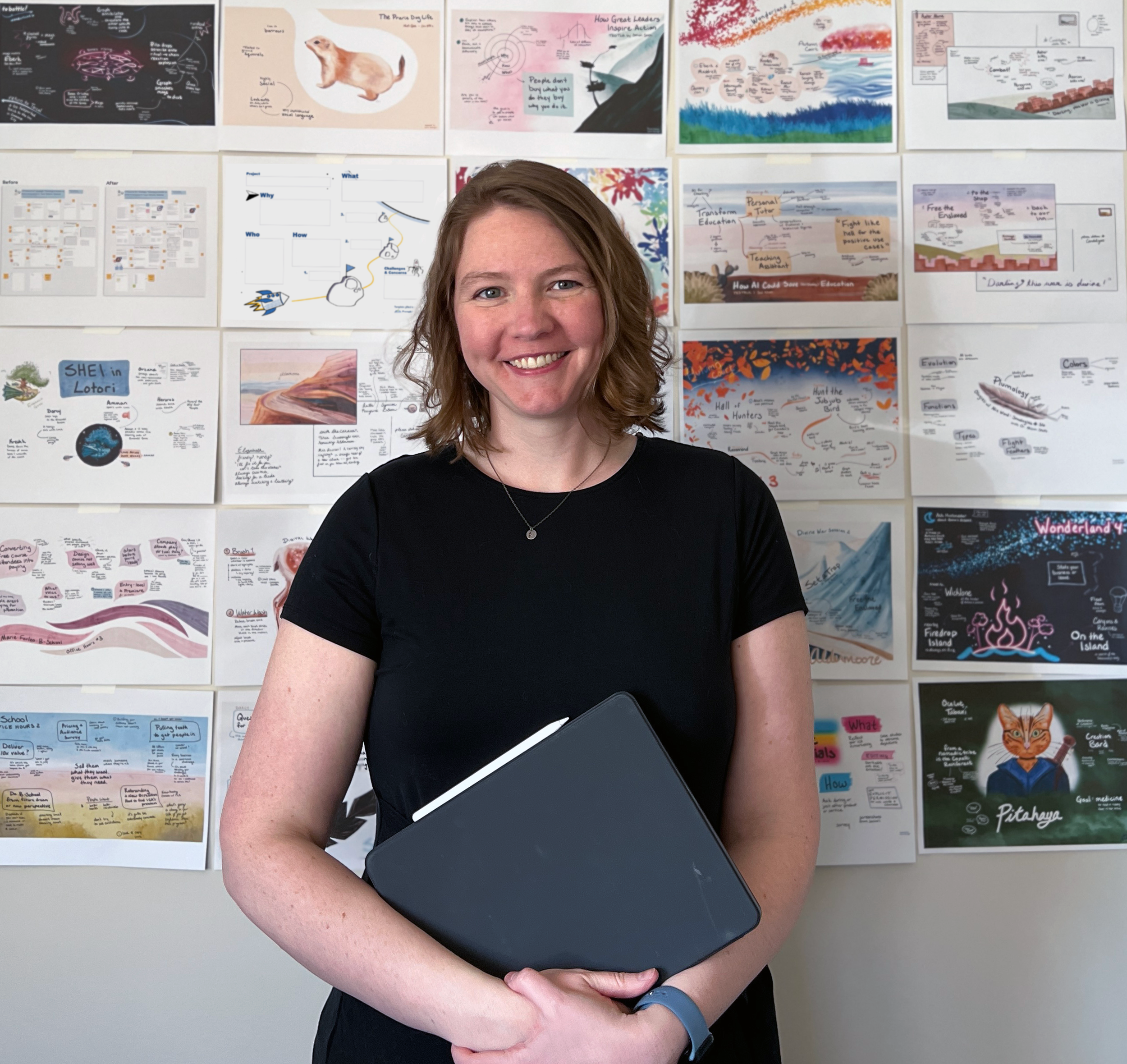 A woman with shoulder-length brown hair, wearing a black T-shirt and a watch, smiling and holding a closed laptop, standing in front of a wall covered with educational posters and illustrations.