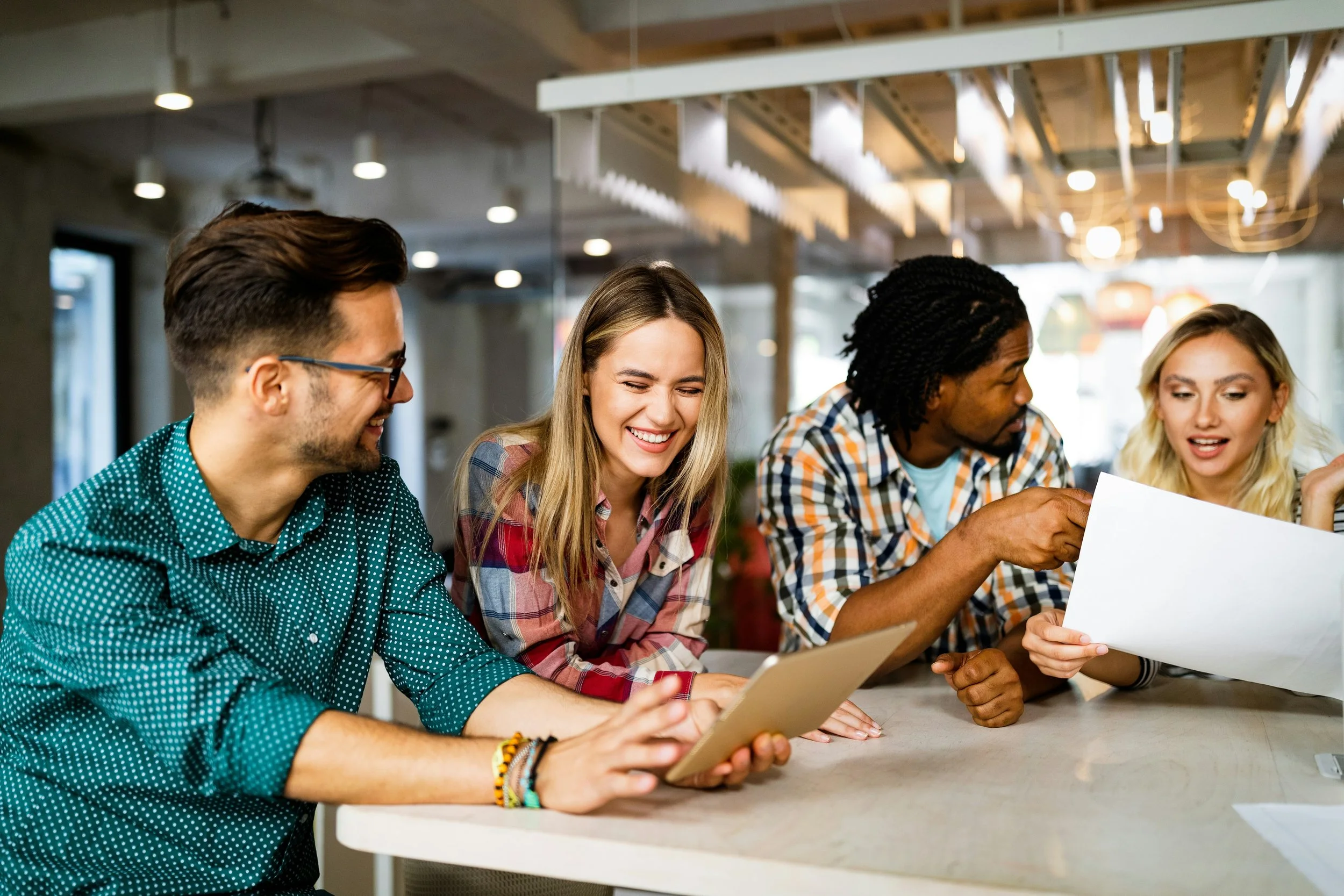 Young adults sitting at a table, smiling and looking at their devices, in a modern indoor setting.