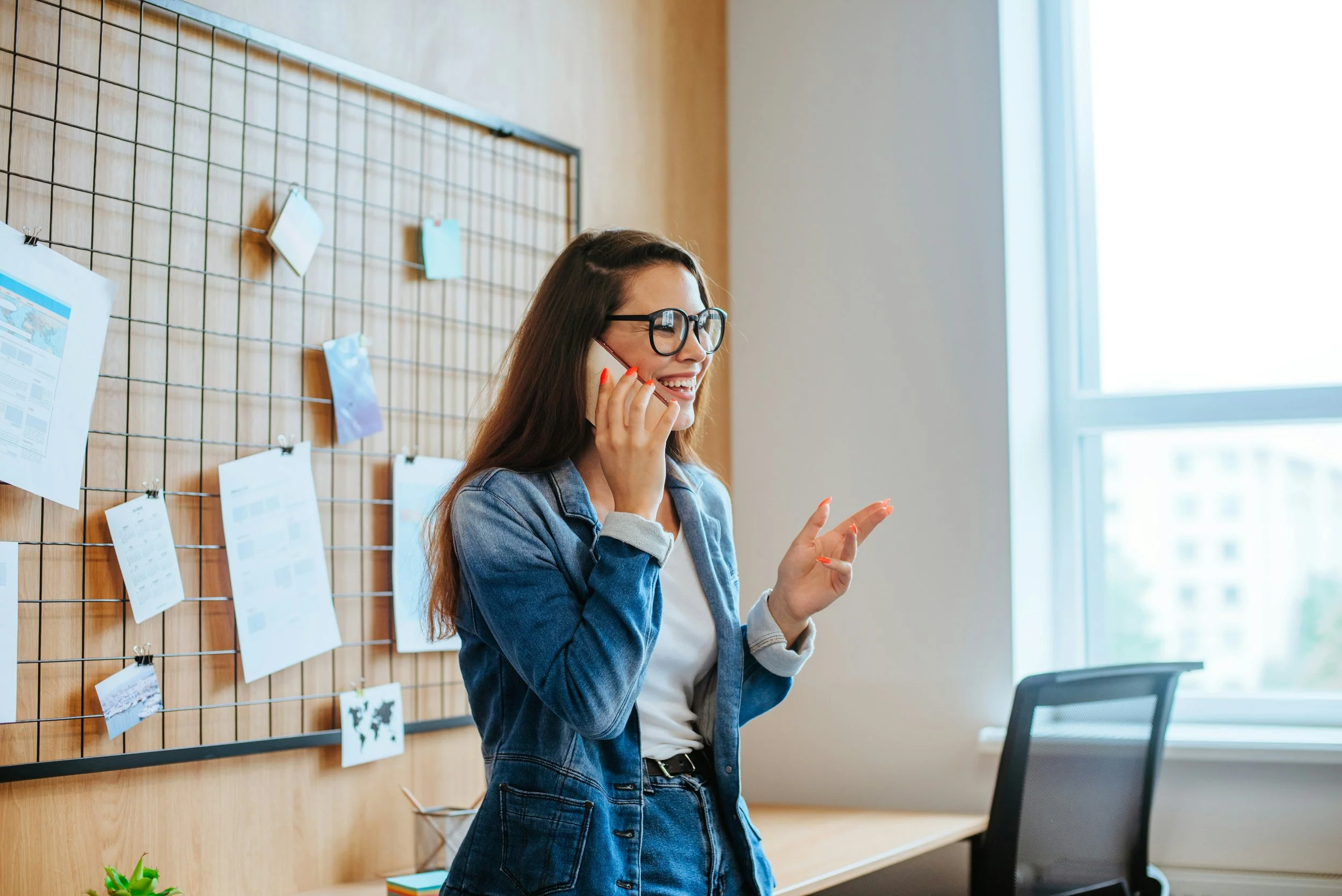 A woman with long dark hair, glasses, wearing a denim jacket, talking on her cell phone and smiling in a modern office with a window and photo display on the wall.
