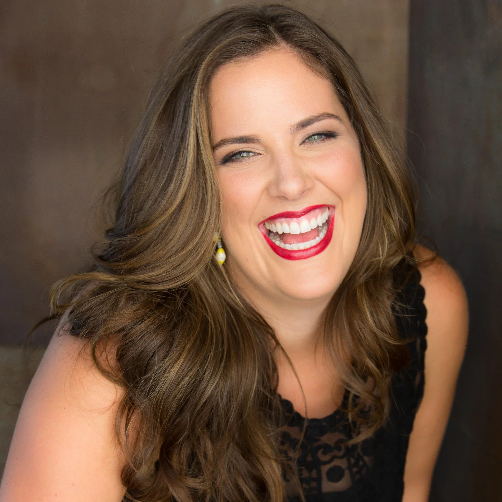 A woman with long wavy brown hair, red lipstick, and a bright smile, wearing a black top with lace details, laughing or smiling enthusiastically.