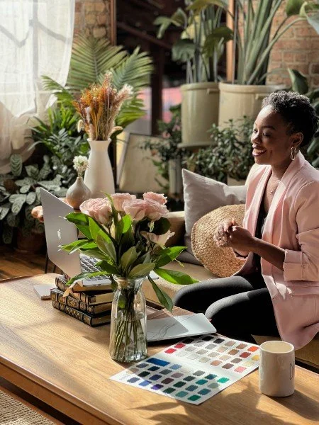 A woman sitting on a couch in a bright, plant-filled room, smiling at her laptop on a wooden coffee table. The table has a large vase of pink roses, a notebook, a coffee mug, and a color palette.