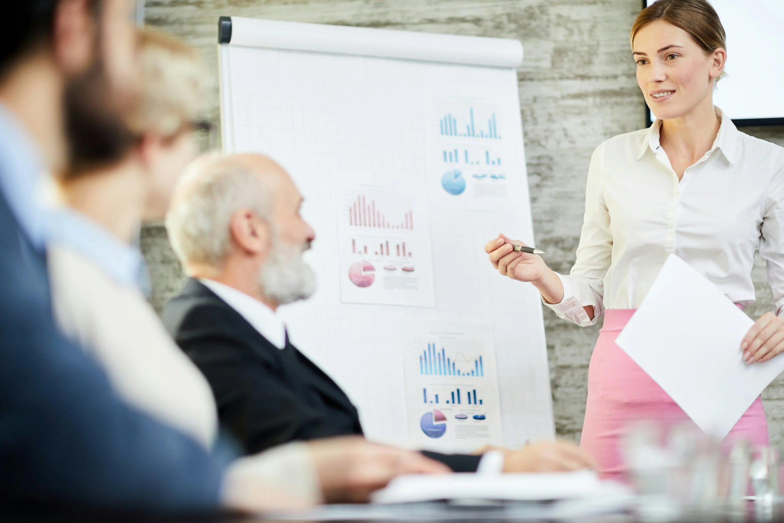 A woman giving a presentation to a group of people during a meeting. She is holding a paper and pointing at a flip chart with charts and graphs.