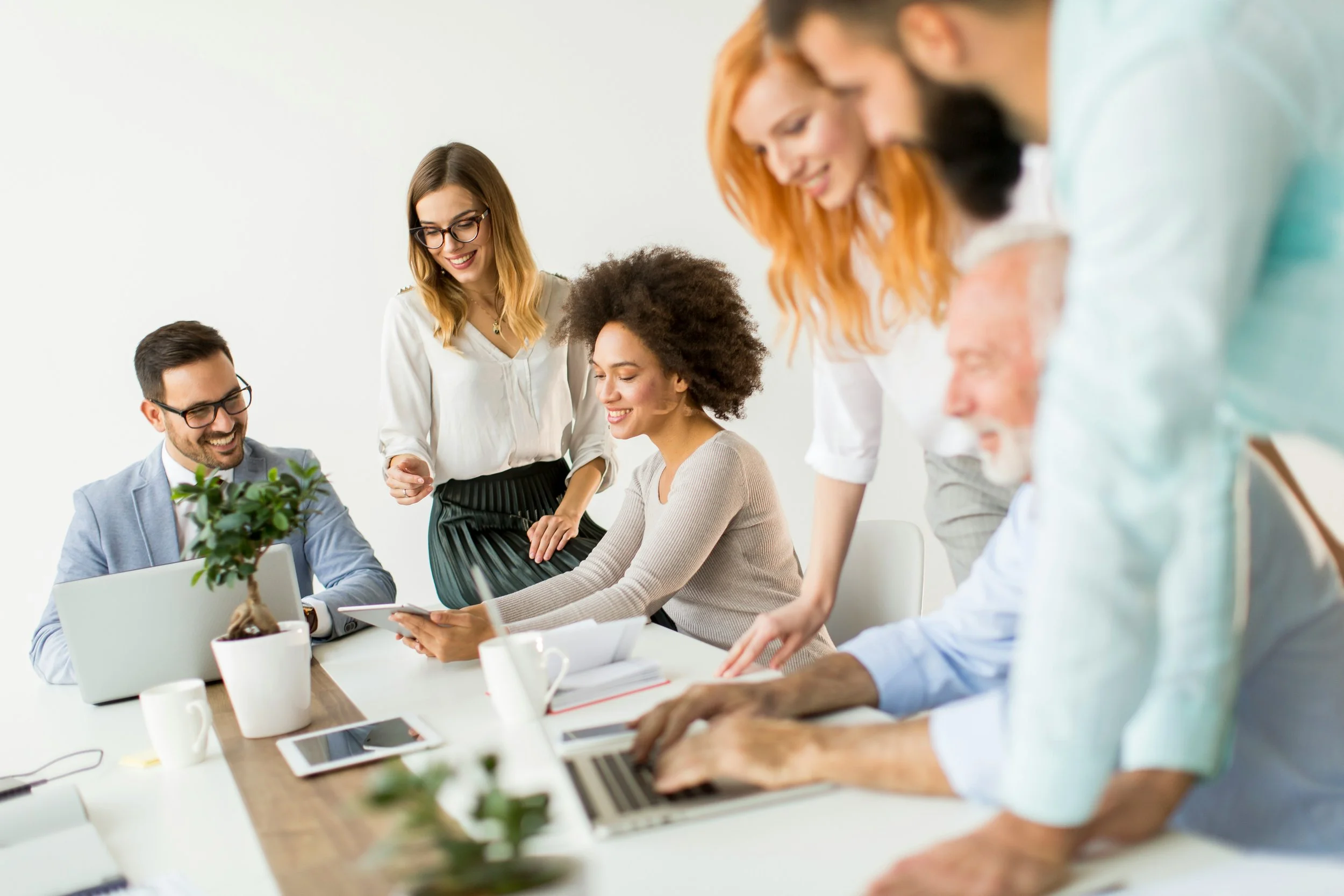 A diverse group of colleagues gathered around a table in a modern office, smiling and collaborating while looking at a tablet and laptops.