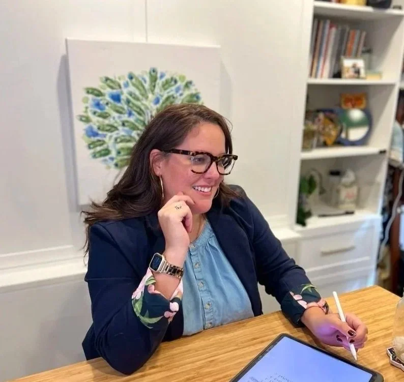 A woman with brown hair, glasses, and a blue top, smiling and holding a white pen, sitting at a wooden table in a cozy room with a white bookshelf and artwork in the background.