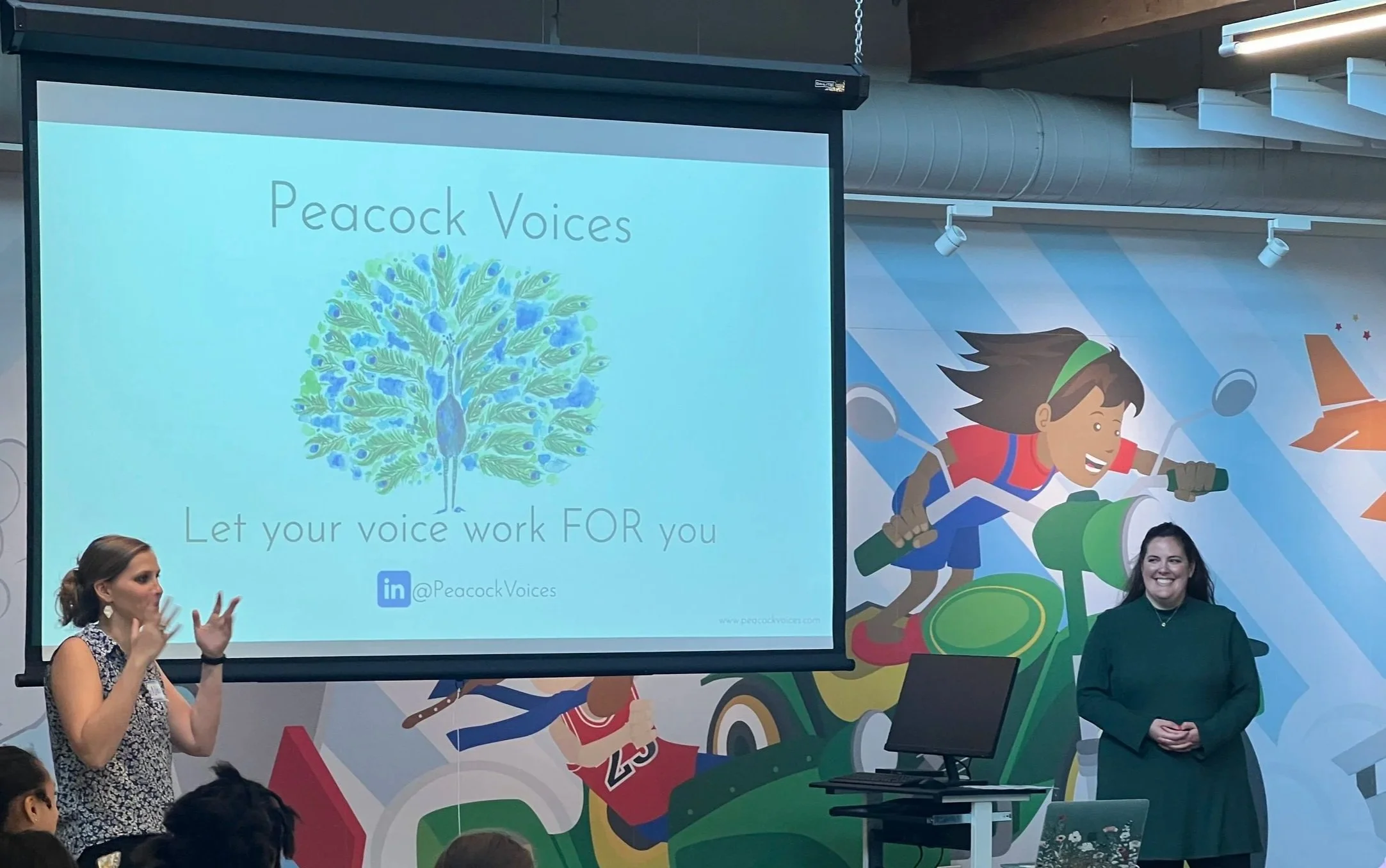 Presentation slide that reads 'Peacock Voices. Let your voice work FOR you' with a peacock illustration. Two women are on stage, one speaking and the other smiling, with a colorful mural of children riding bikes and airplanes in the background.