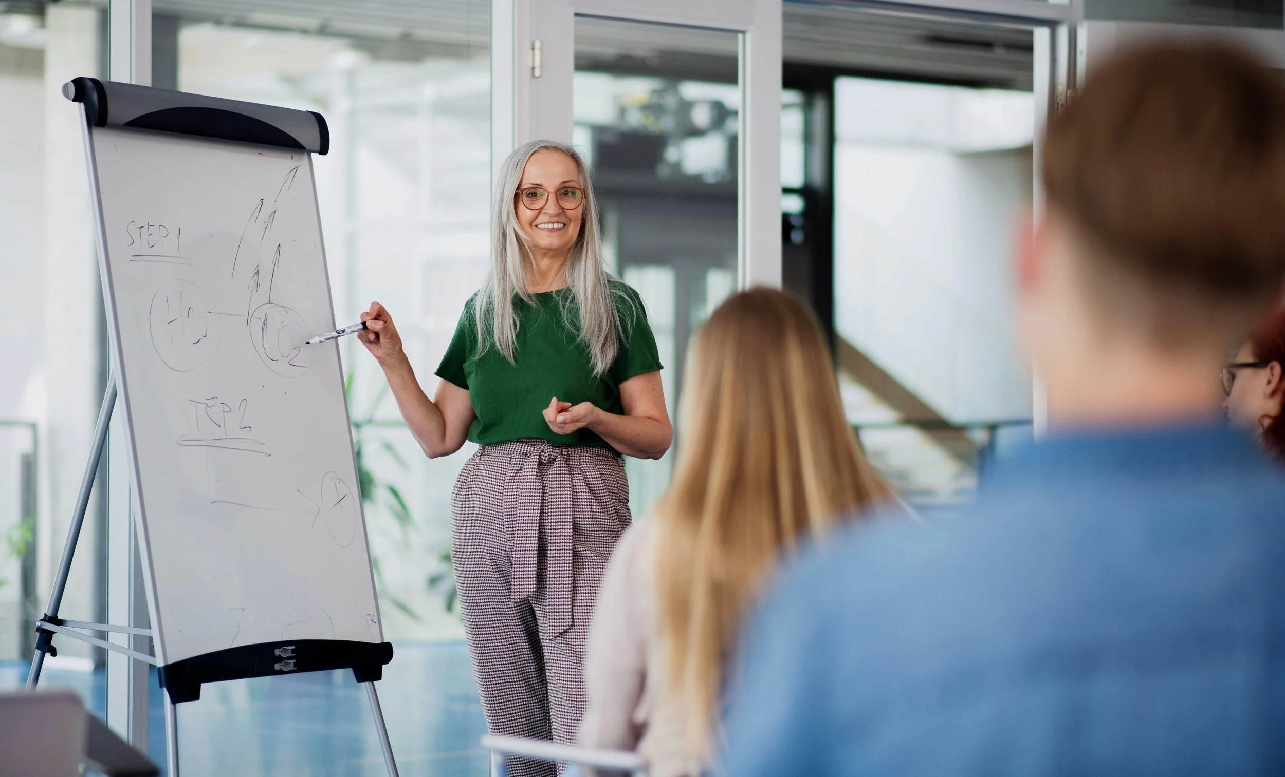 A woman with long gray hair, glasses, and a green top is standing next to a whiteboard, giving a presentation to a group of people in an office setting. The whiteboard has diagrams and the word "Step 1" written on it, with a marker in her hand. The audience is listening attentively.