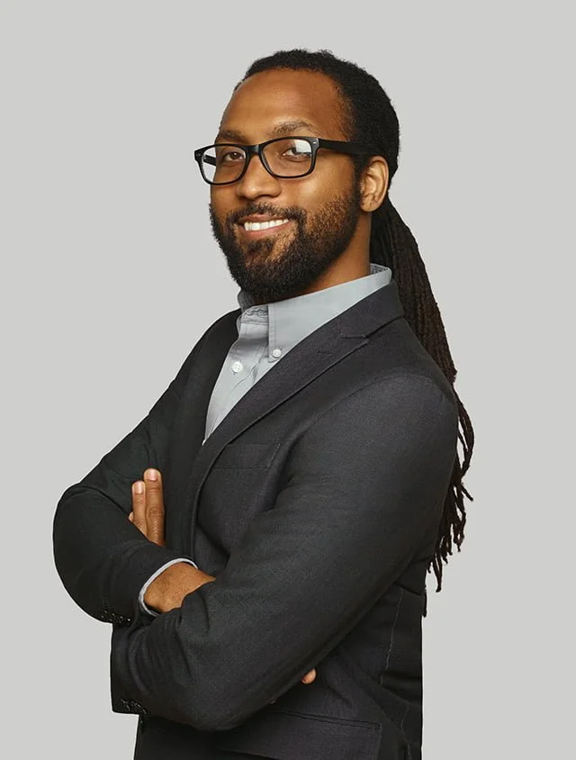 A man with long dreadlocks, beard, and glasses, wearing a dark suit jacket and a light shirt, smiling with arms crossed against a plain gray background.