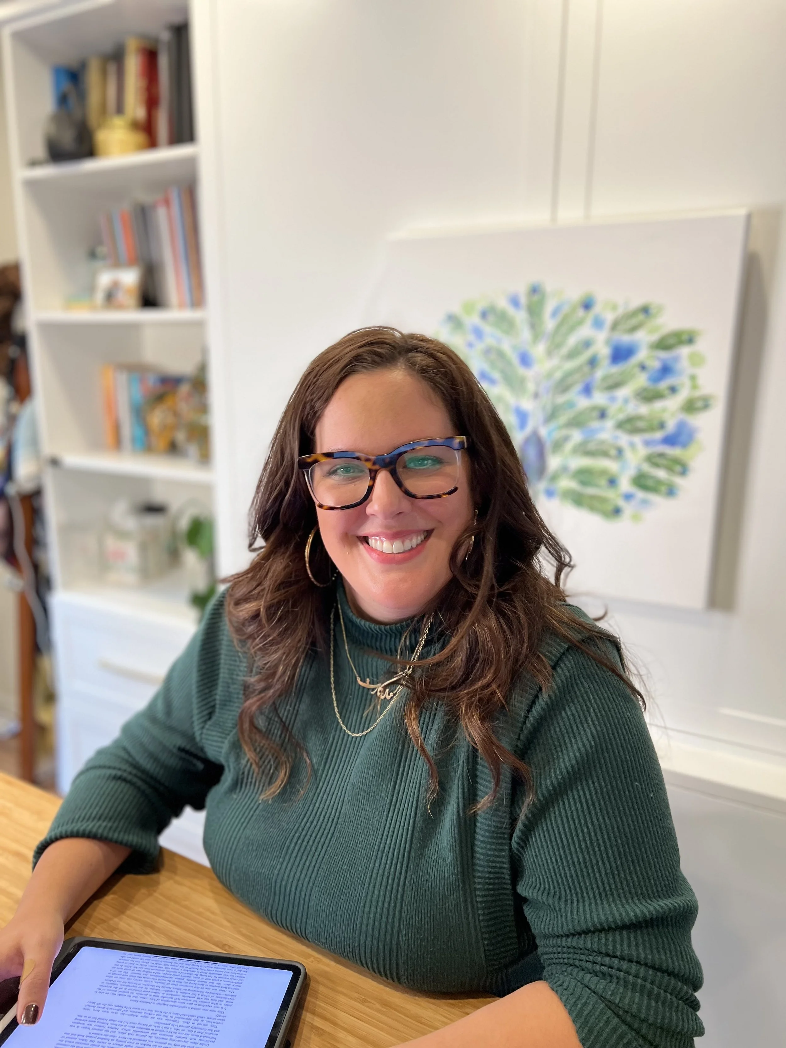 A woman with brown hair, wearing glasses, gold hoop earrings, a green ribbed sweater, and layered gold necklaces, sitting at a wooden table with a tablet in front of her. She is smiling and looking at the camera. In the background, there is a white bookshelf with books and decorations and a colorful abstract painting of a tree.