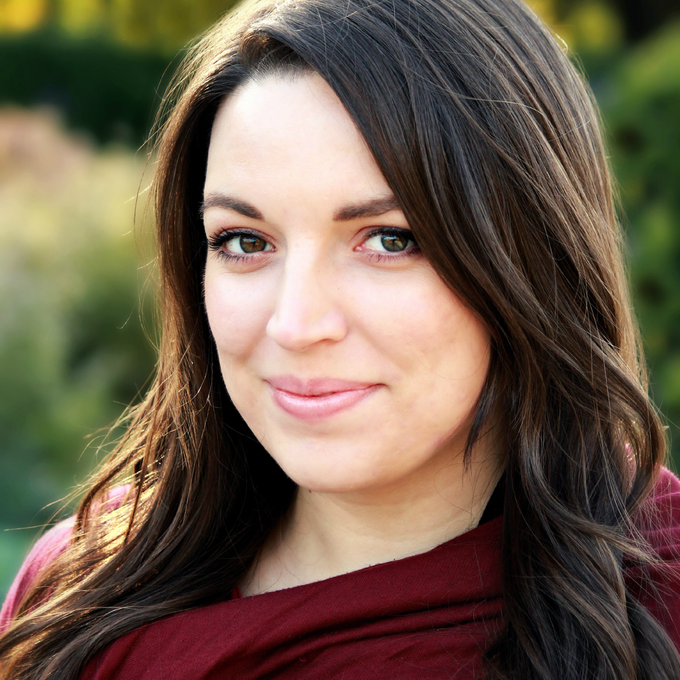 Close-up portrait of a young woman with long brown hair and blue eyes, wearing a maroon top outdoors with blurred greenery in the background.