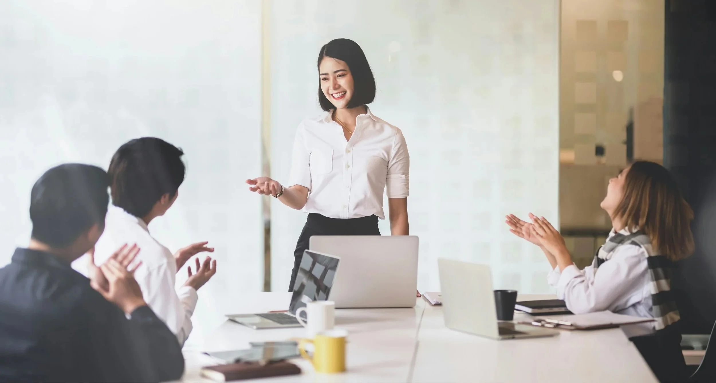 A woman in a white shirt standing and smiling in a meeting room, performing a presentation while four seated colleagues clap and smile.