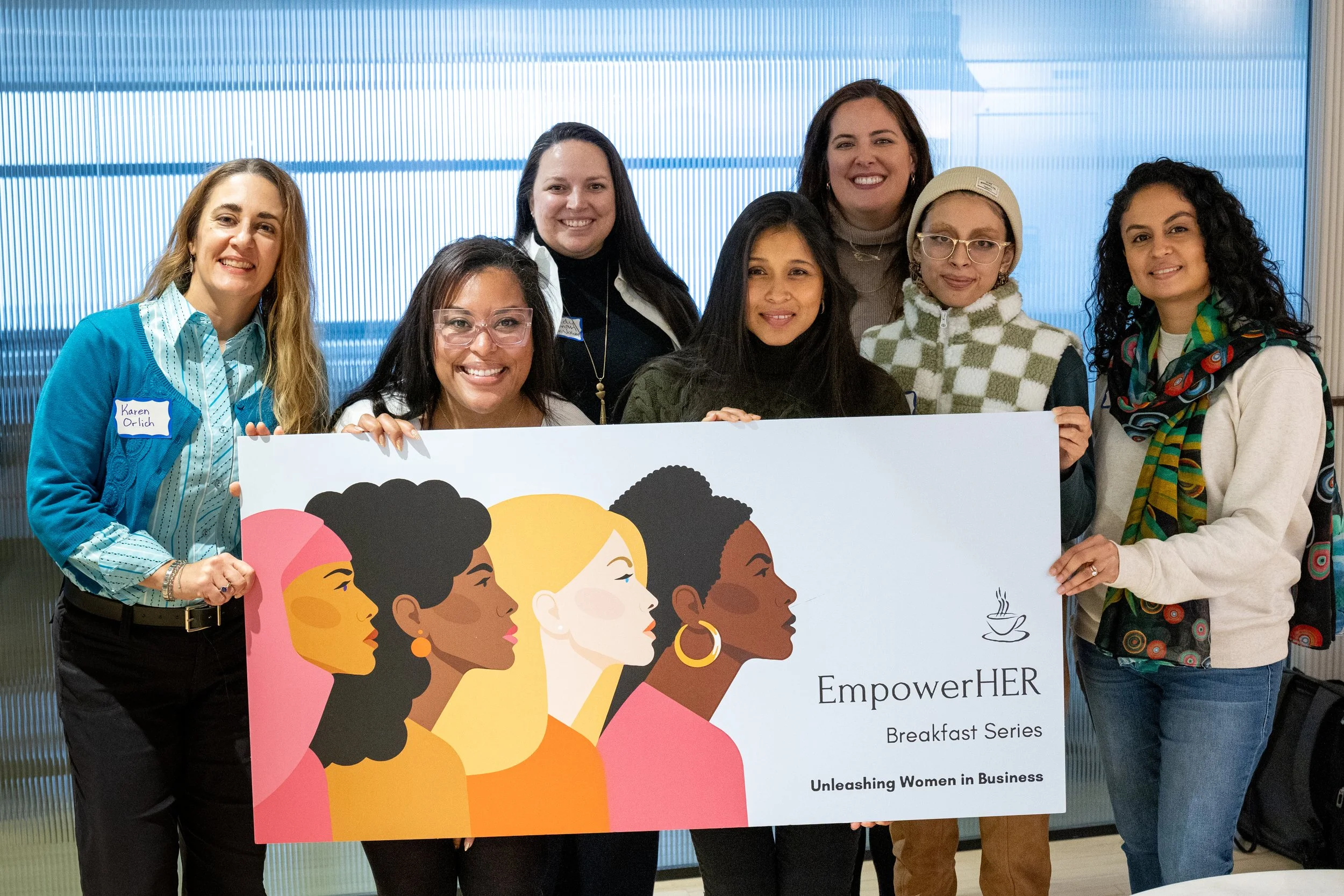 Group of women smiling, holding a sign that says "EmpowerHER Breakfast Series: Unleashing Women in Business", with a colorful illustration of diverse women.