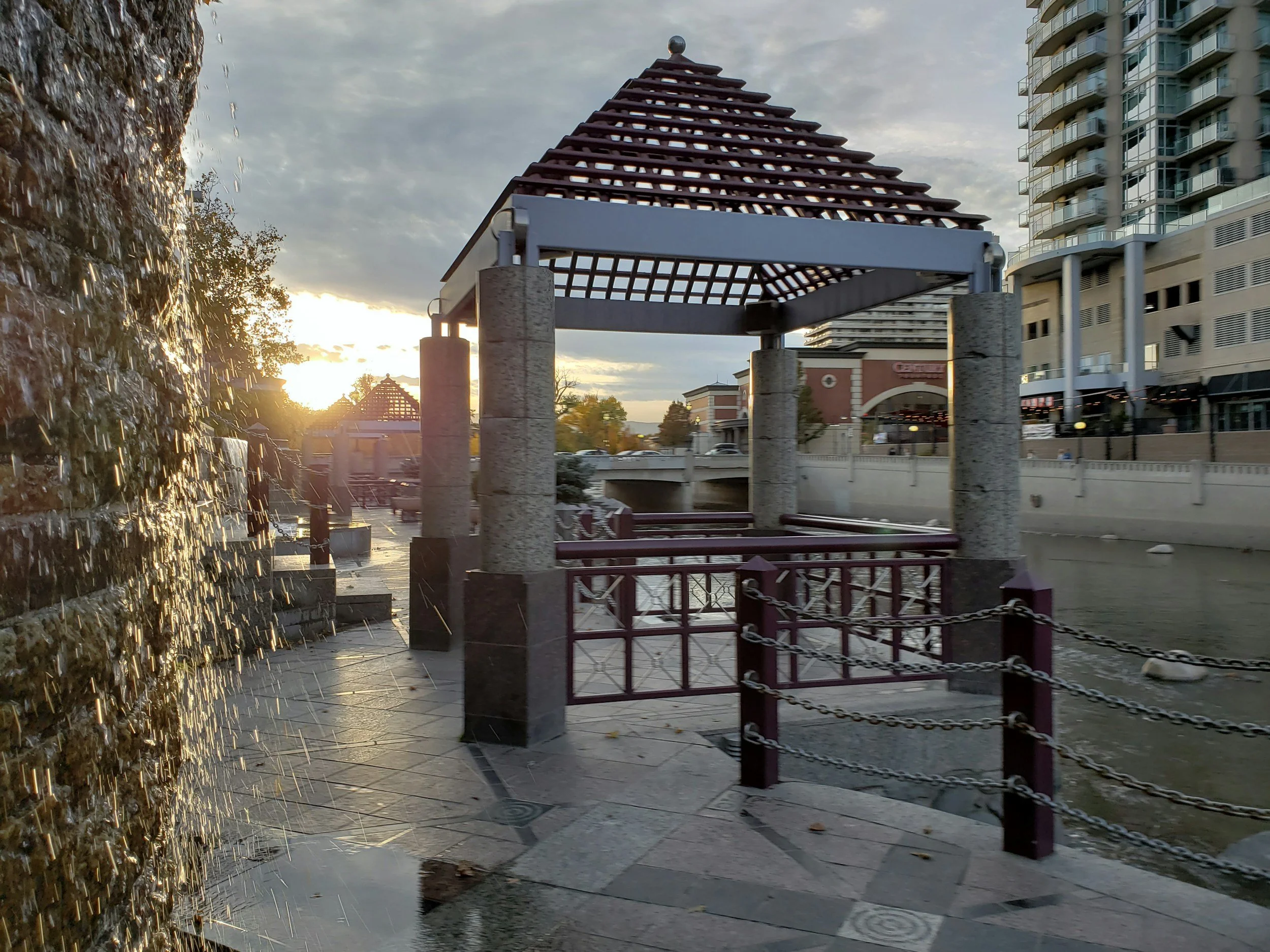 A waterfront park scene during sunset in Reno with a waterfall feature on the left, a decorative pavilion with a wooden lattice roof, and modern buildings in the background.