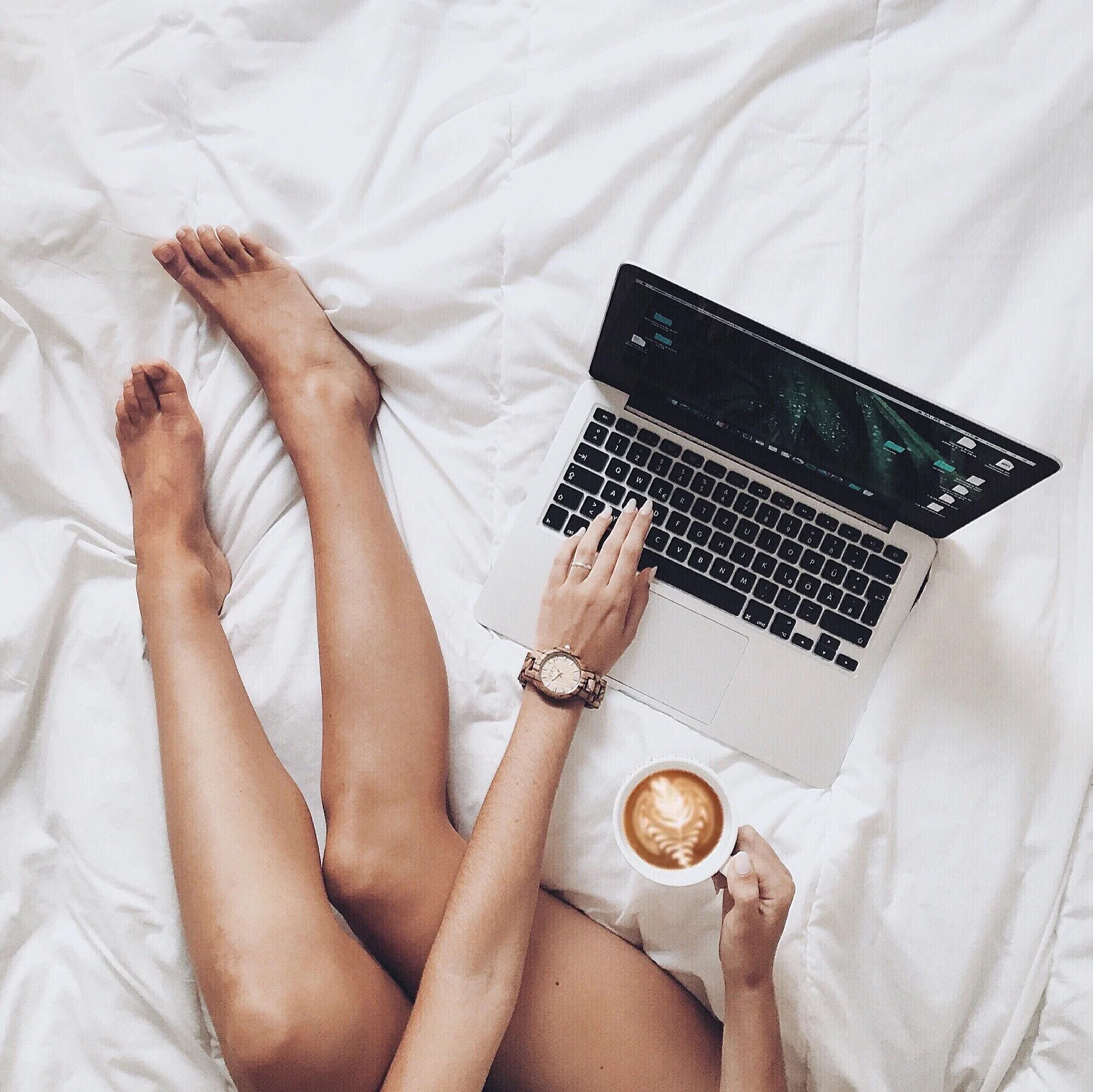 A person sitting on a bed with white sheets, holding a cup of coffee with latte art in one hand and using a laptop with the other. The person is wearing a large watch on their wrist.