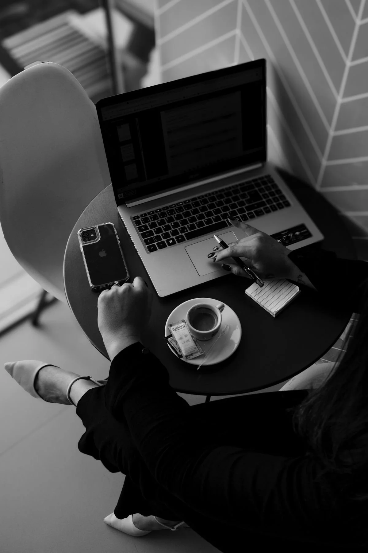 A person sitting at a round table working on a laptop, with a smartphone, a cup of coffee, and a small notepad on the table. The person is wearing high heels and has long hair.