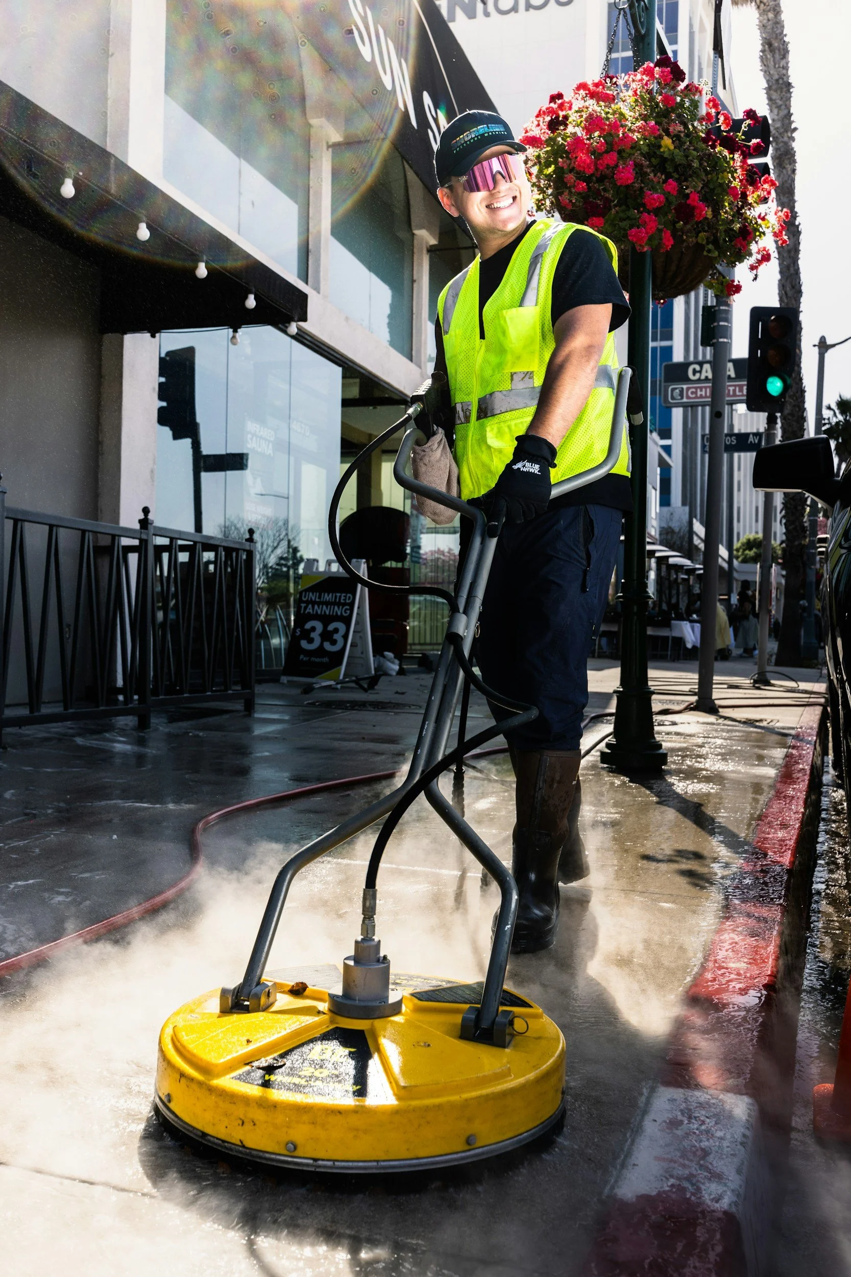 A street worker wearing a yellow safety vest, goggles, and gloves uses a power street cleaner with yellow base to spray water on the sidewalk, with a building and a sign advertising tanning in the background.