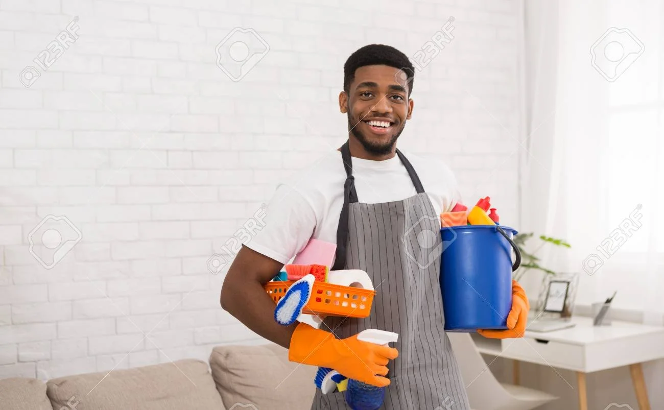 Smiling man wearing gray apron holding cleaning supplies in a bright living room.