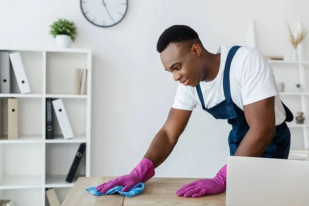 A young man cleaning a wooden table with a cloth, wearing pink gloves and a white T-shirt with blue overalls in a bright, organized room with shelves, books, and a plant.