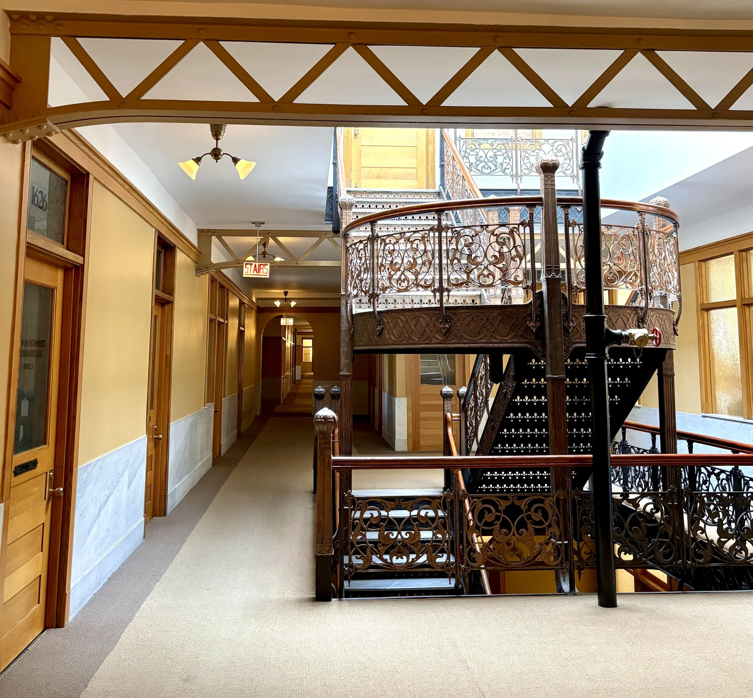 Interior view of a corridor in a historic building with wooden doors, yellow walls, and a decorative wrought iron staircase.