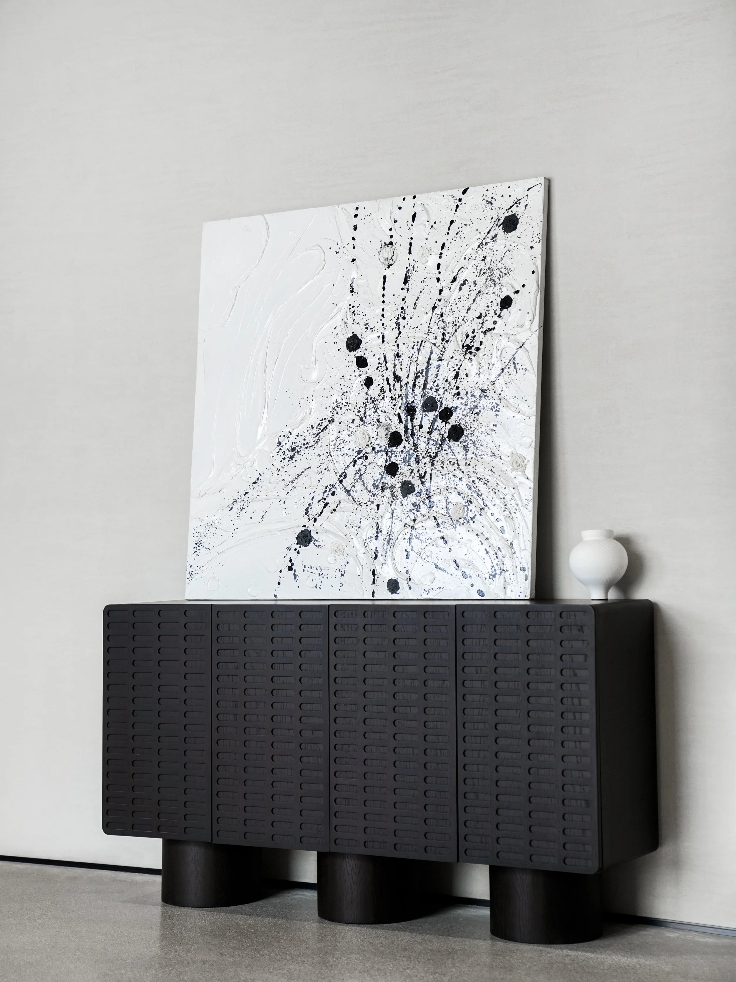 Abstract black and white splatter painting on canvas resting on a black cabinet with a white vase on top, against a light-colored wall.