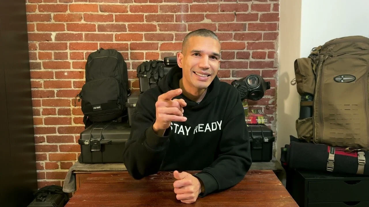 Clark at his studio desk wearing a Stay Ready hoodie, with preparedness equipment behind him.