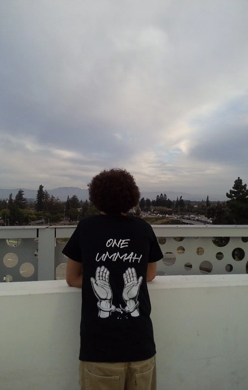 Person with curly hair facing away, wearing a black T-shirt with 'One Ummah' and two raised hands graphic, standing on a balcony overlooking trees, buildings, and a cloudy sky in the distance.