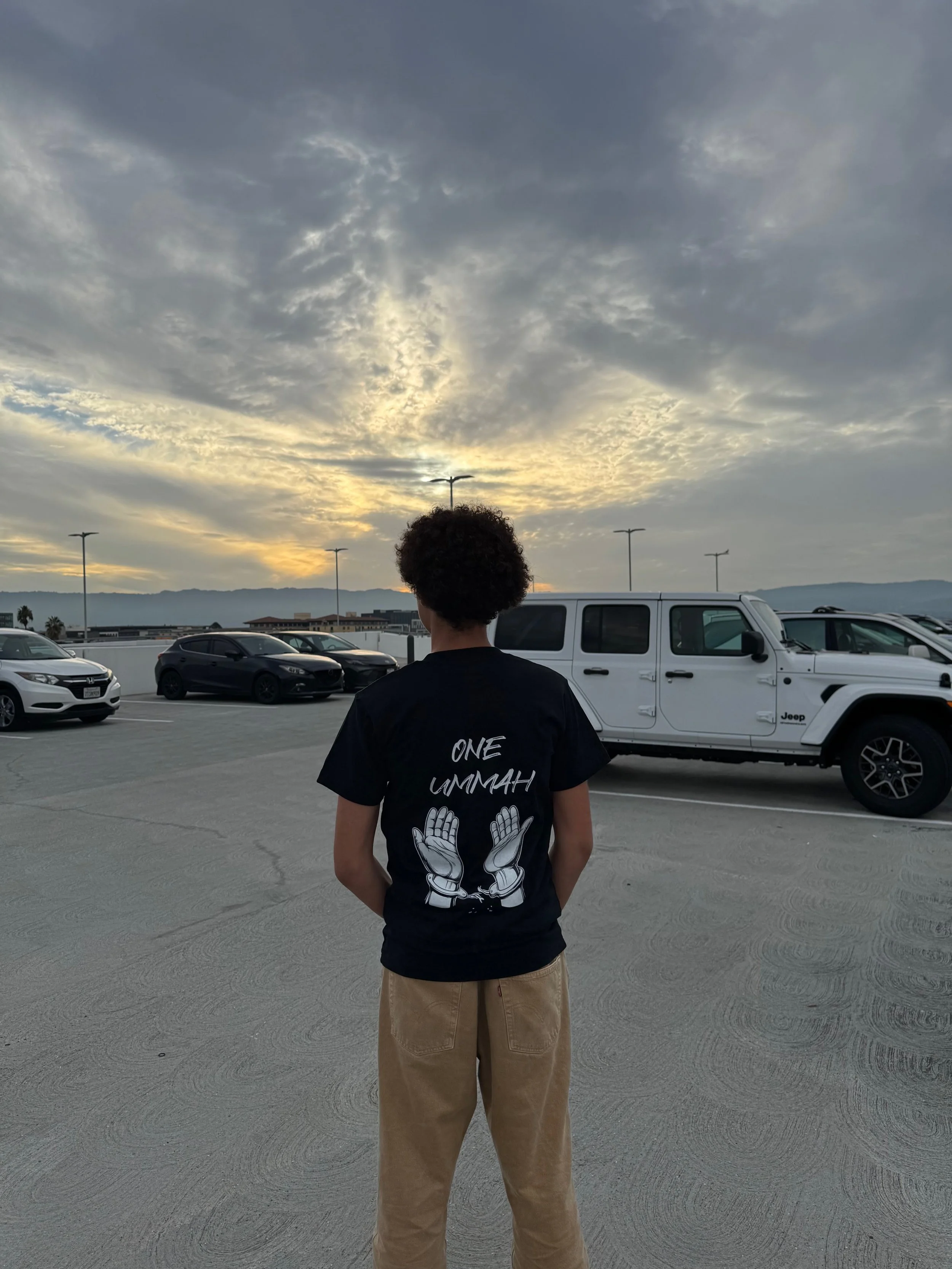 Person standing in a parking lot during sunset, wearing a black t-shirt with 'ONE LMMAH' and praying hands graphic, beige pants, parked cars, and a cloudy sky.