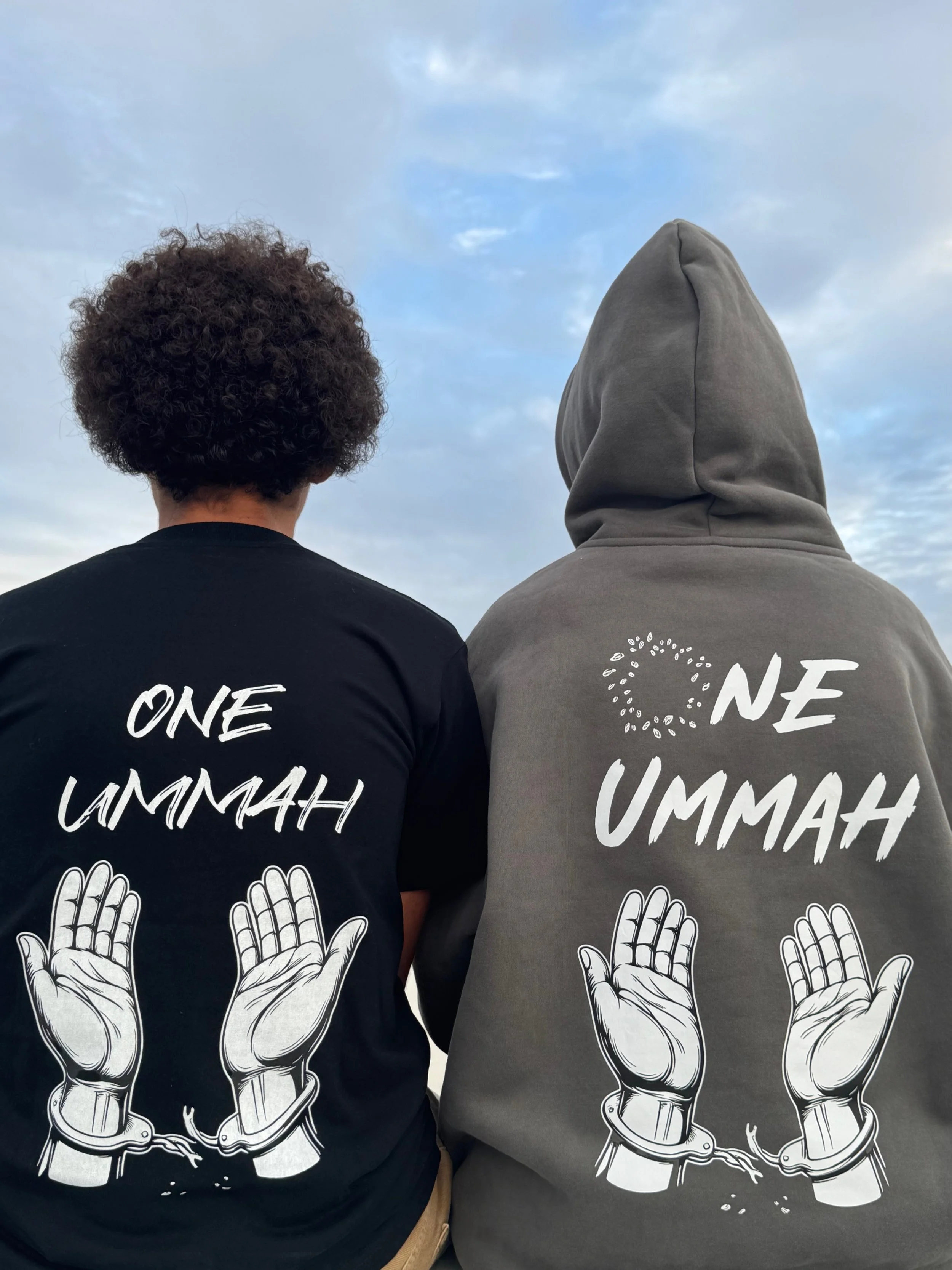Two people sitting side by side wearing hoodies and T-shirts with the message "One Ummah" and illustrations of broken handcuffs, symbolizing unity and freedom, with a cloudy sky in the background.