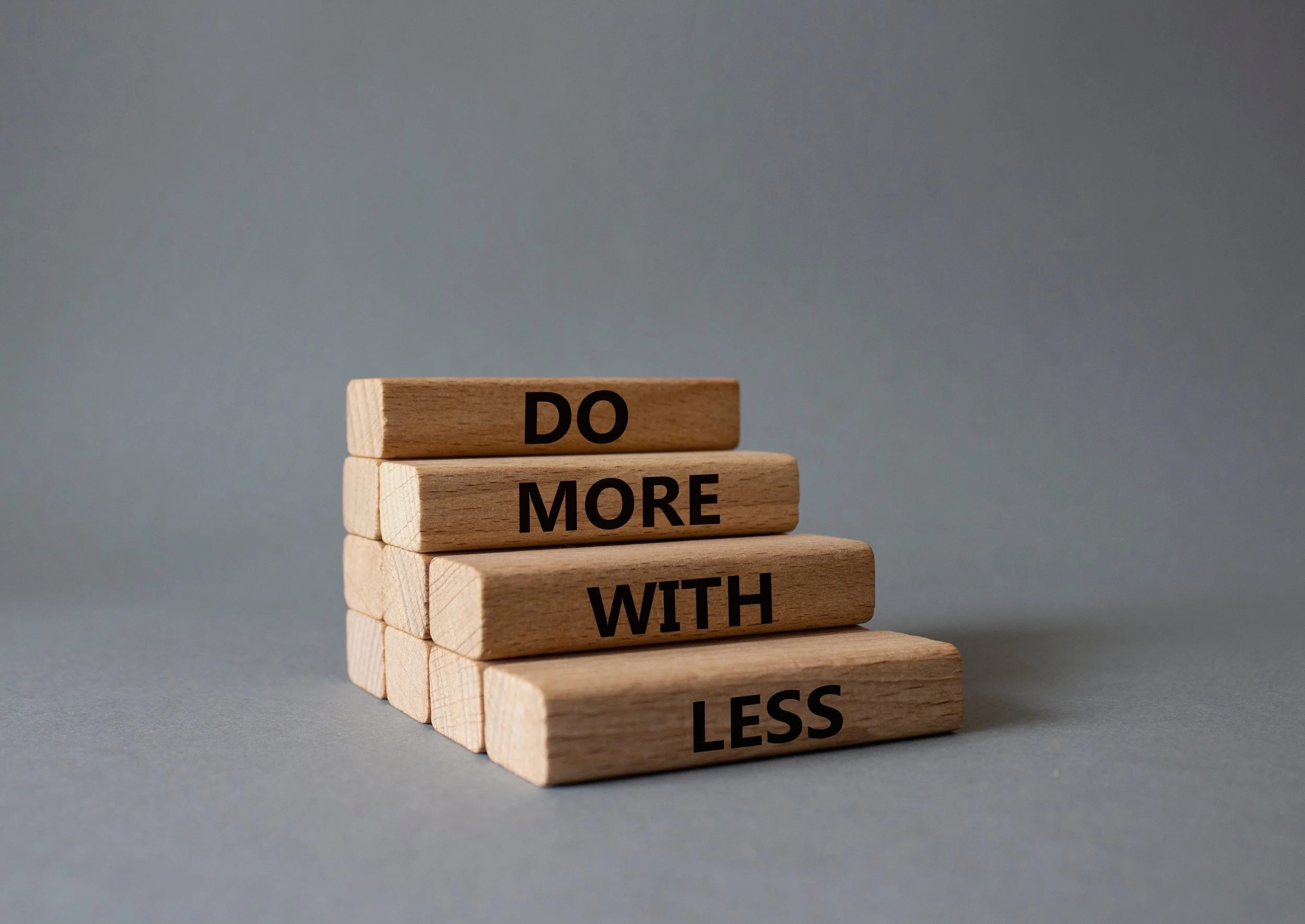 Stacked wooden blocks with words 'DO MORE WITH LESS' printed in black, arranged in increasing height from bottom to top on a gray surface with a plain gray background.