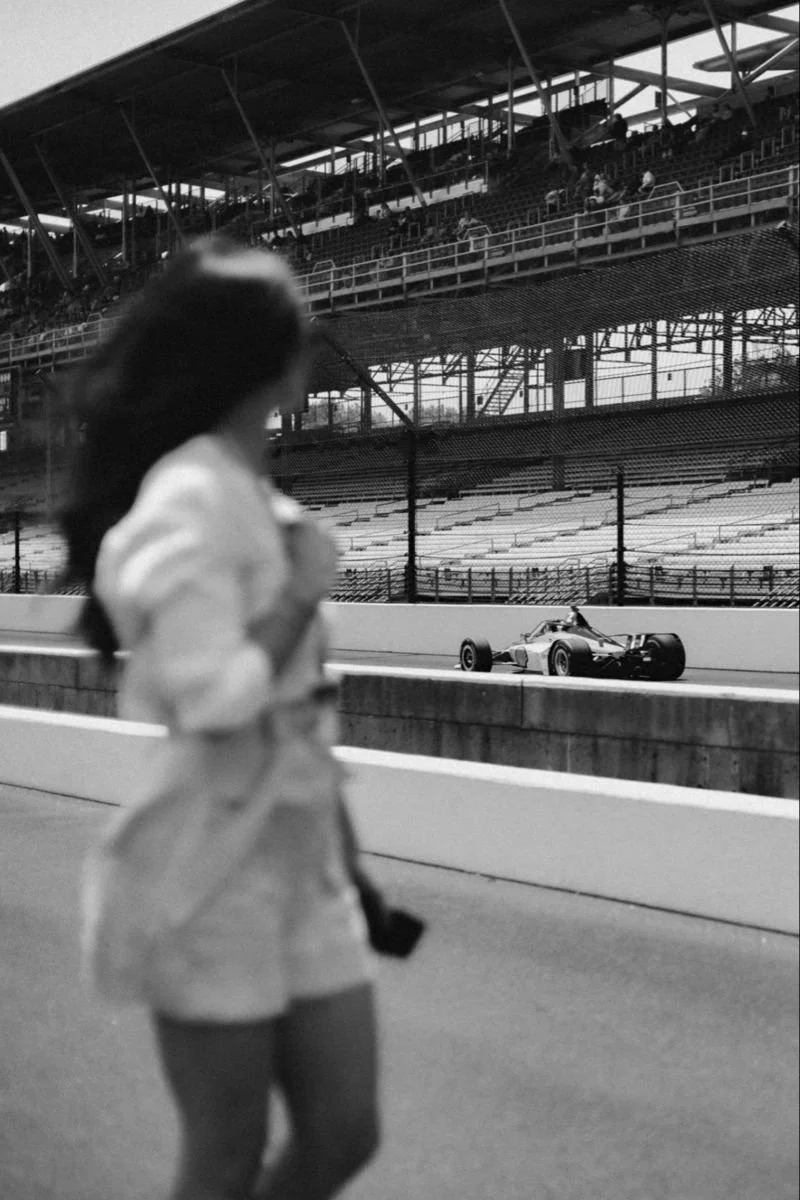 A woman in a light-colored jacket and shorts standing at a race track, with a race car passing by on the track and empty spectator stands in the background.