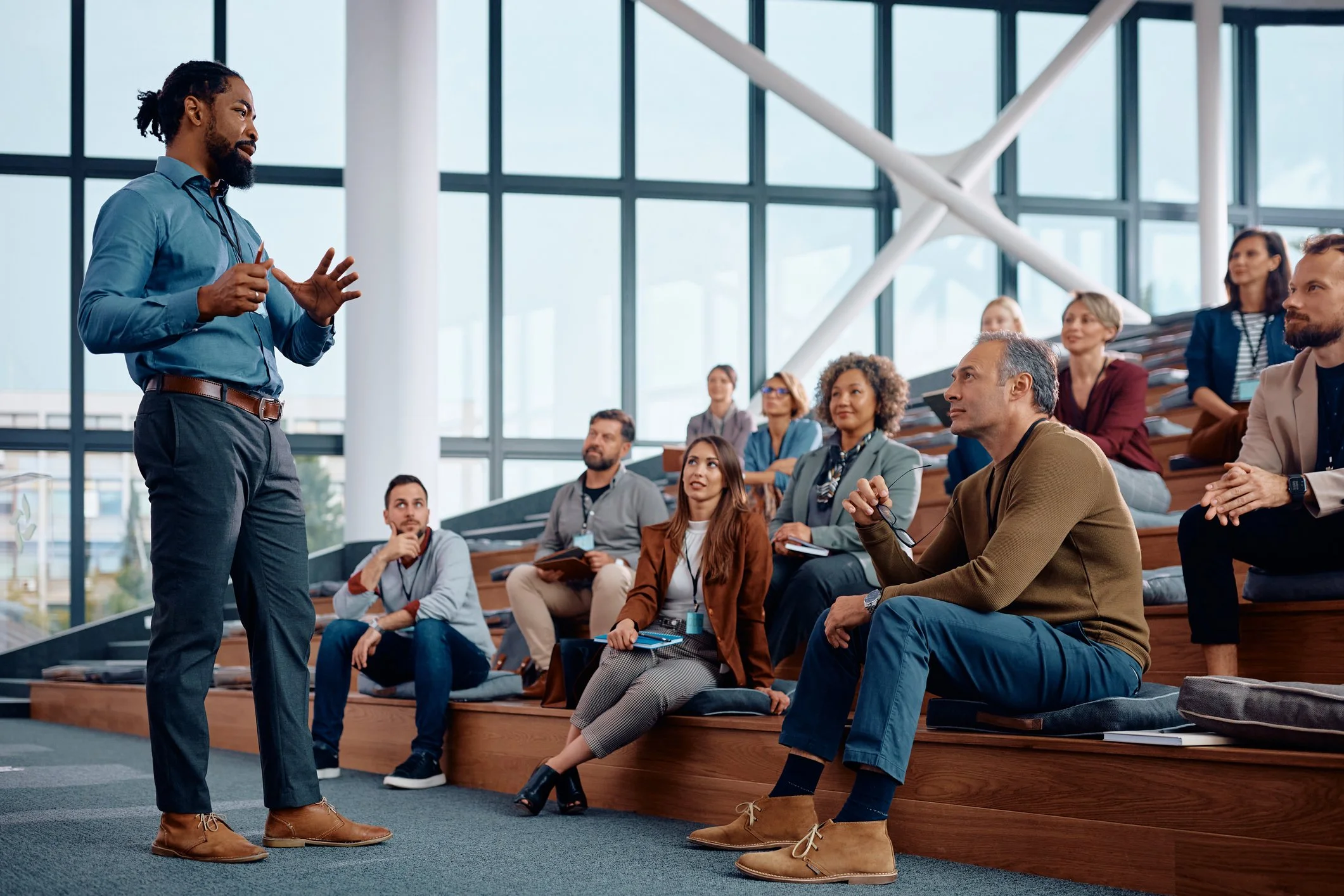 A man with dreadlocks giving a presentation to an audience seated on tiered wooden benches inside a modern conference room with large glass windows.