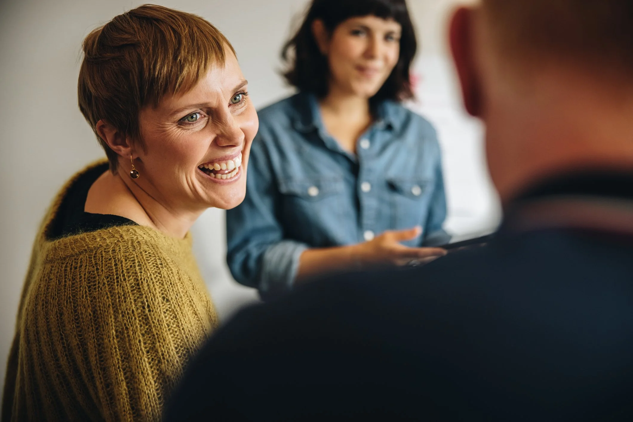 Three people engaged in conversation, with a woman smiling and talking, a woman in the background holding a tablet, and part of a man's shoulder visible in the foreground.