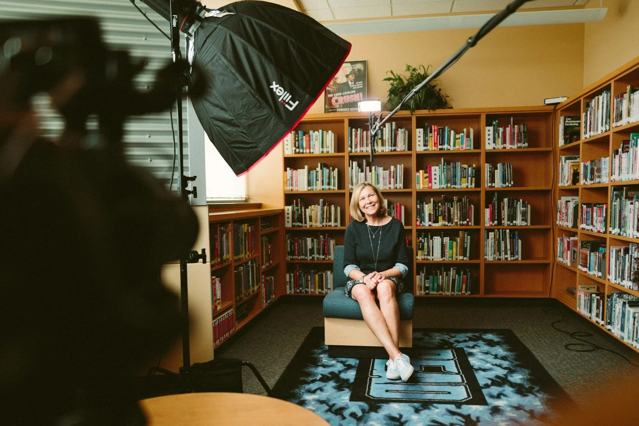 A woman smiling while sitting on a chair in front of bookshelves in a library during a video recording or interview, with lighting equipment visible.