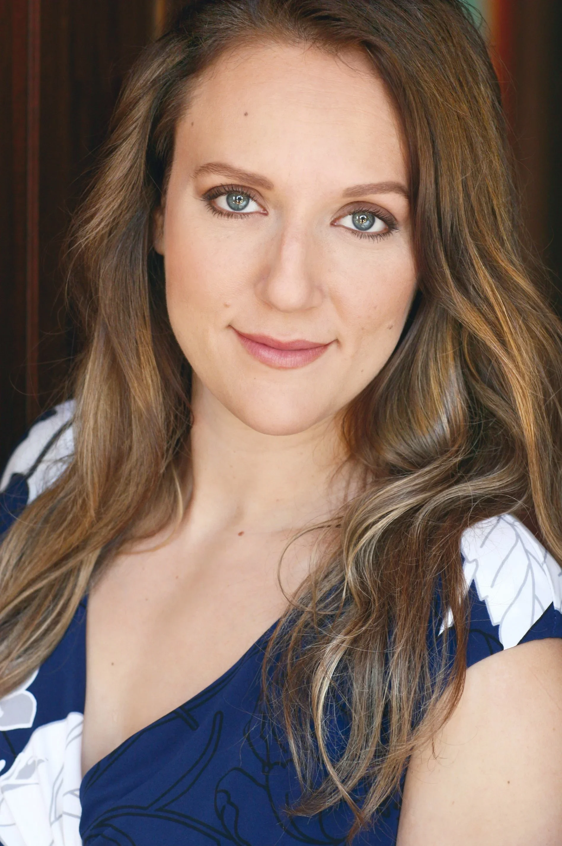 Close-up portrait of a woman with blue eyes and long wavy brown hair, wearing a blue and white floral dress, smiling softly at the camera.