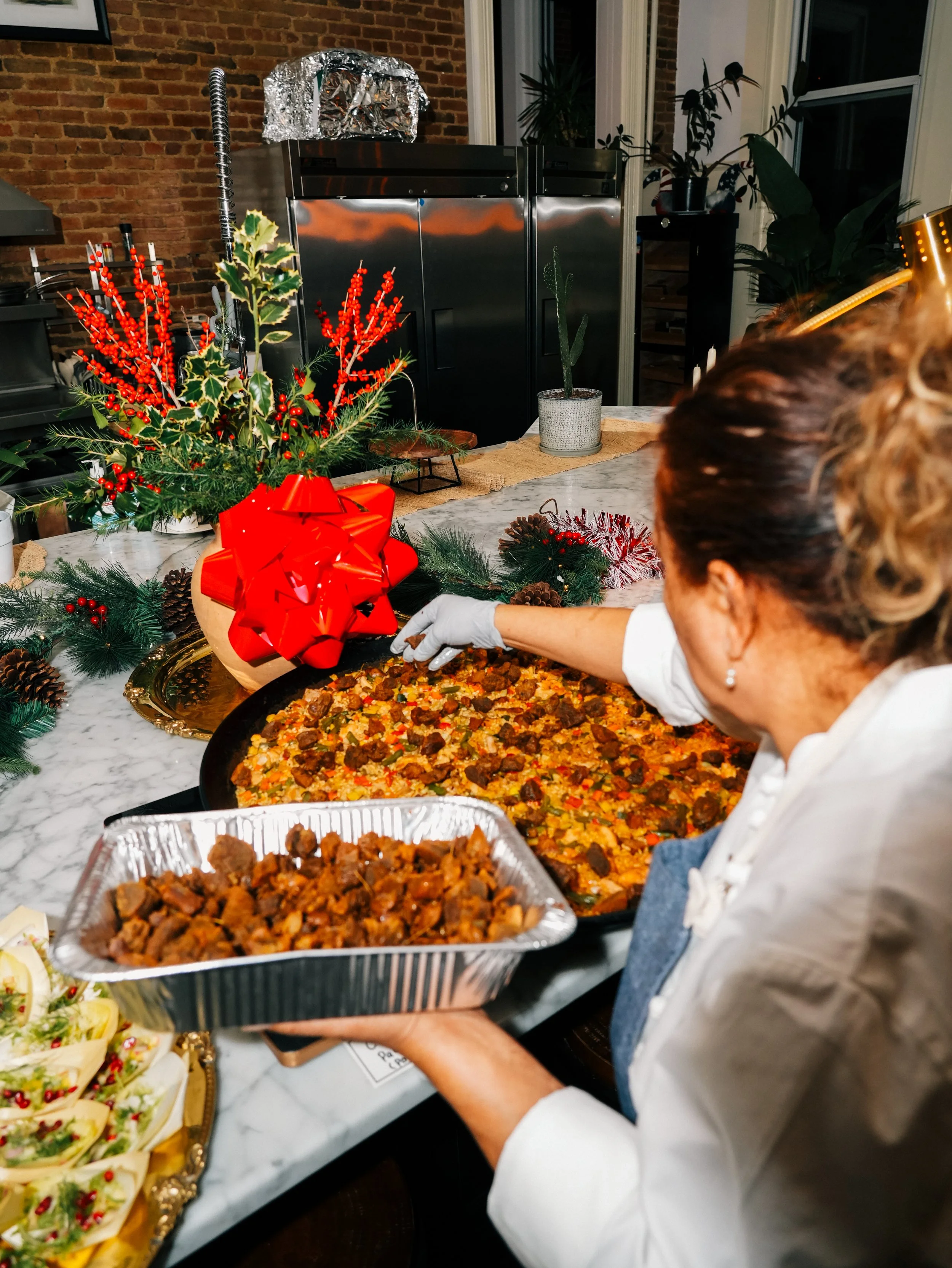 A woman wearing a white shirt and apron serving a large dish of cooked food at a festive holiday table decorated with greenery, pinecones, and a red bow, with a Christmas-themed floral arrangement and other dishes on the table.