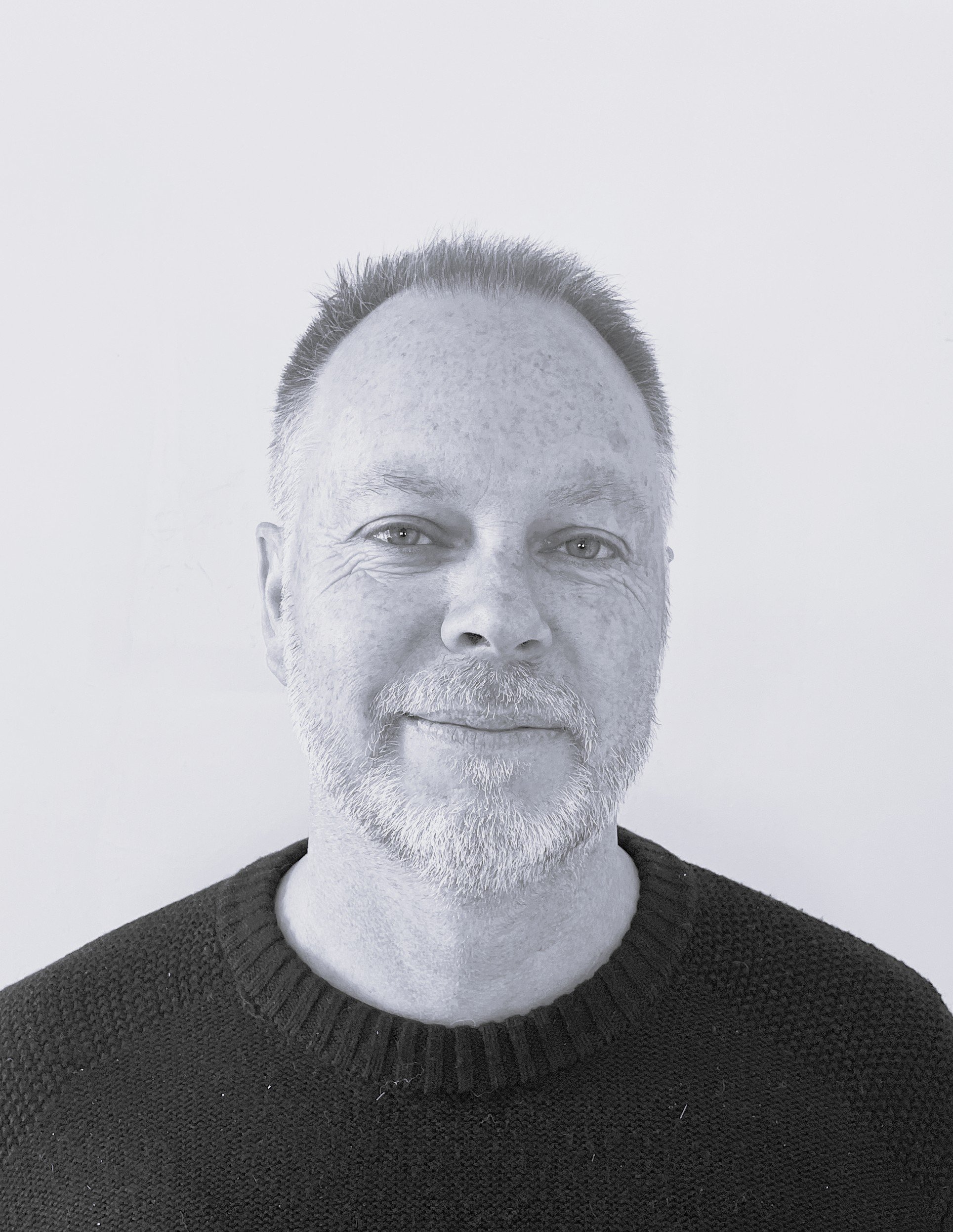A black and white portrait of a middle-aged man with short hair, a beard, and light-colored eyes, wearing a dark knit sweater against a plain background.