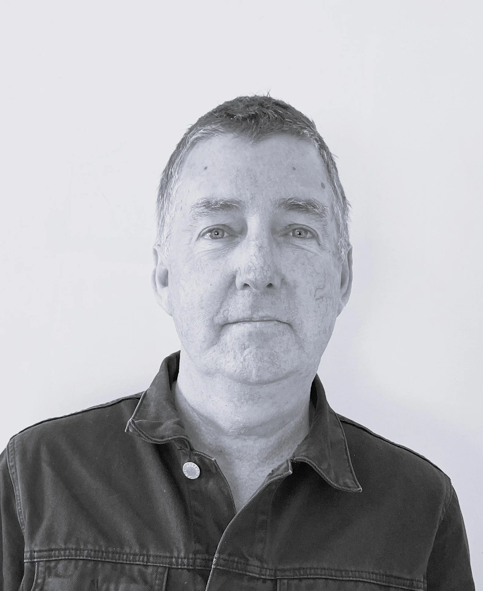 Black-and-white photo of a man with short, dark hair wearing a dark jacket standing against a plain white background.