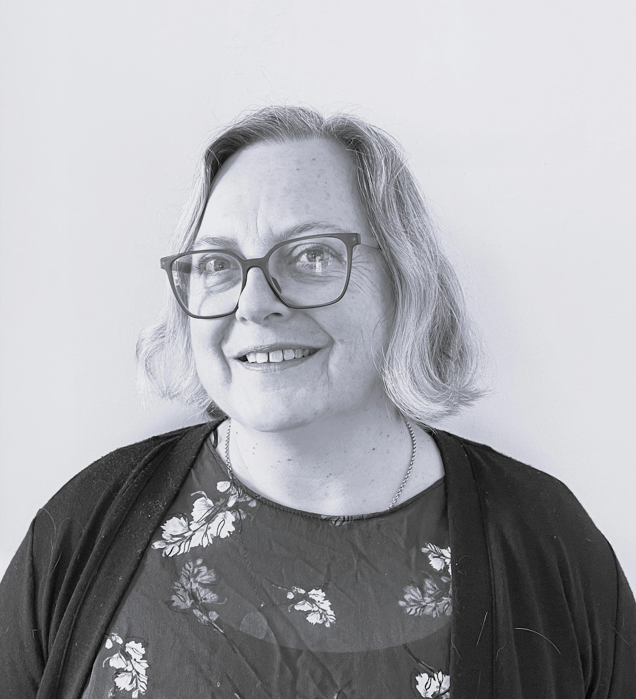 Black and white portrait of a smiling woman with short hair and glasses, wearing a floral blouse and a necklace, standing against a plain background.