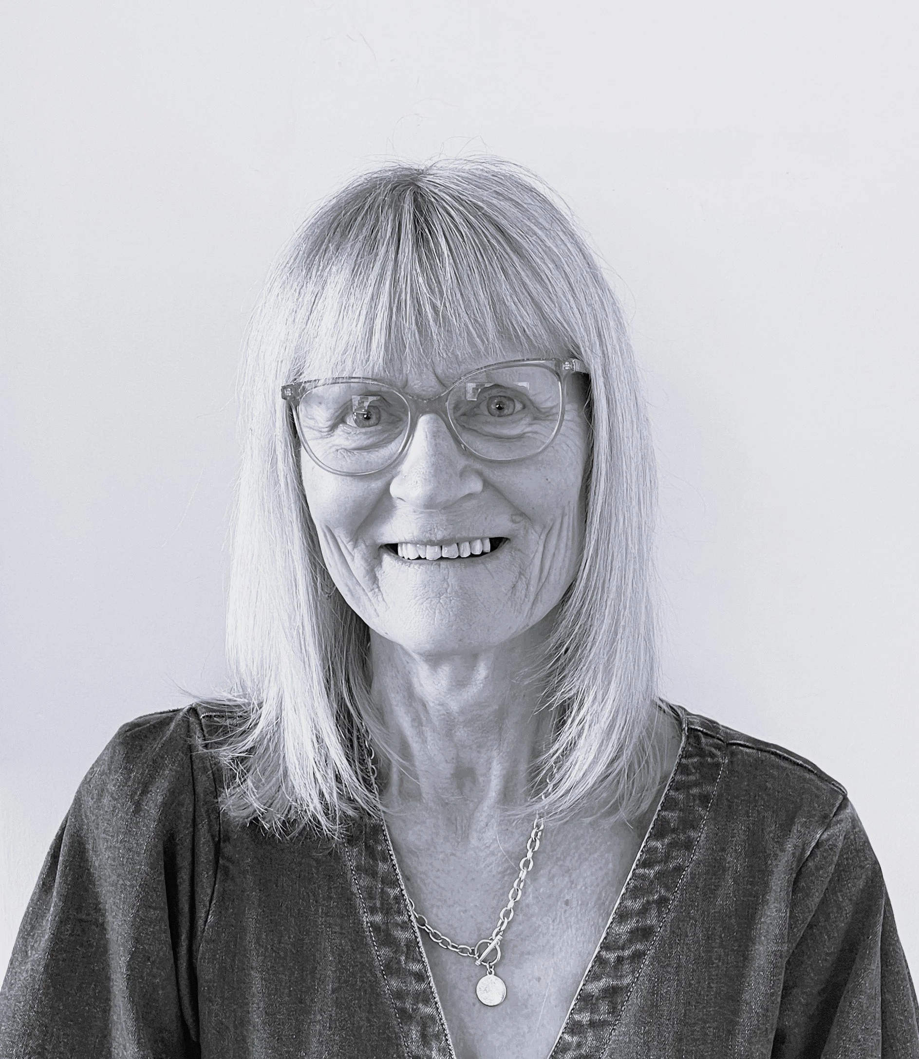 Black and white photo of an elderly woman with shoulder-length hair, glasses, and a necklace, smiling.