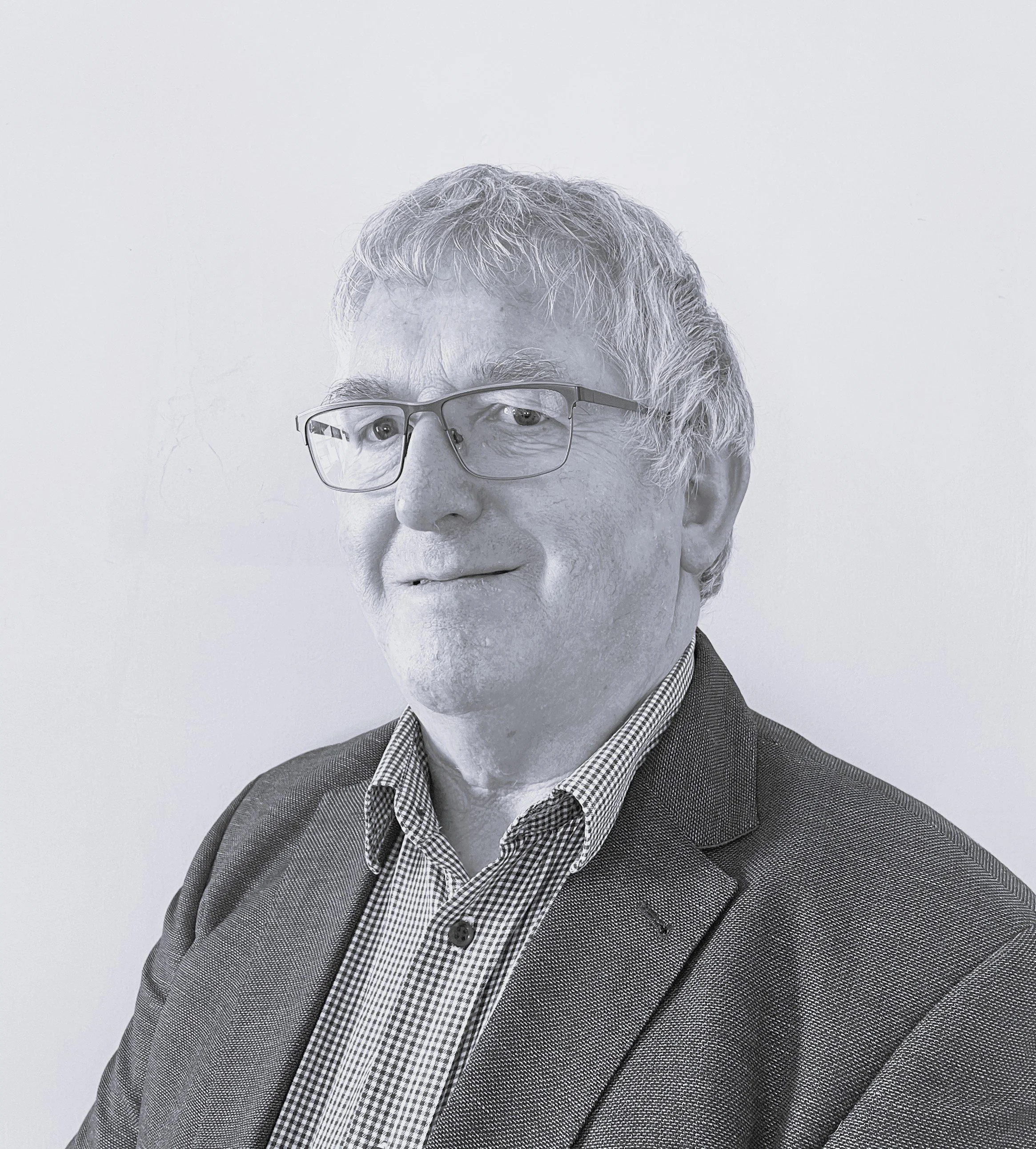 A black-and-white portrait of an older man with glasses, light-colored hair, wearing a houndstooth shirt and a blazer, smiling subtly against a plain background.