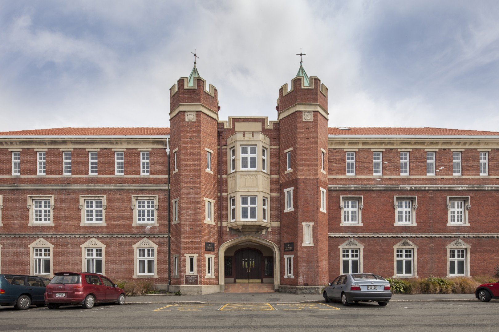 Front view of a red brick building with castle-like features, including two round towers and an arched entrance, parked cars in front, and a cloudy sky above.