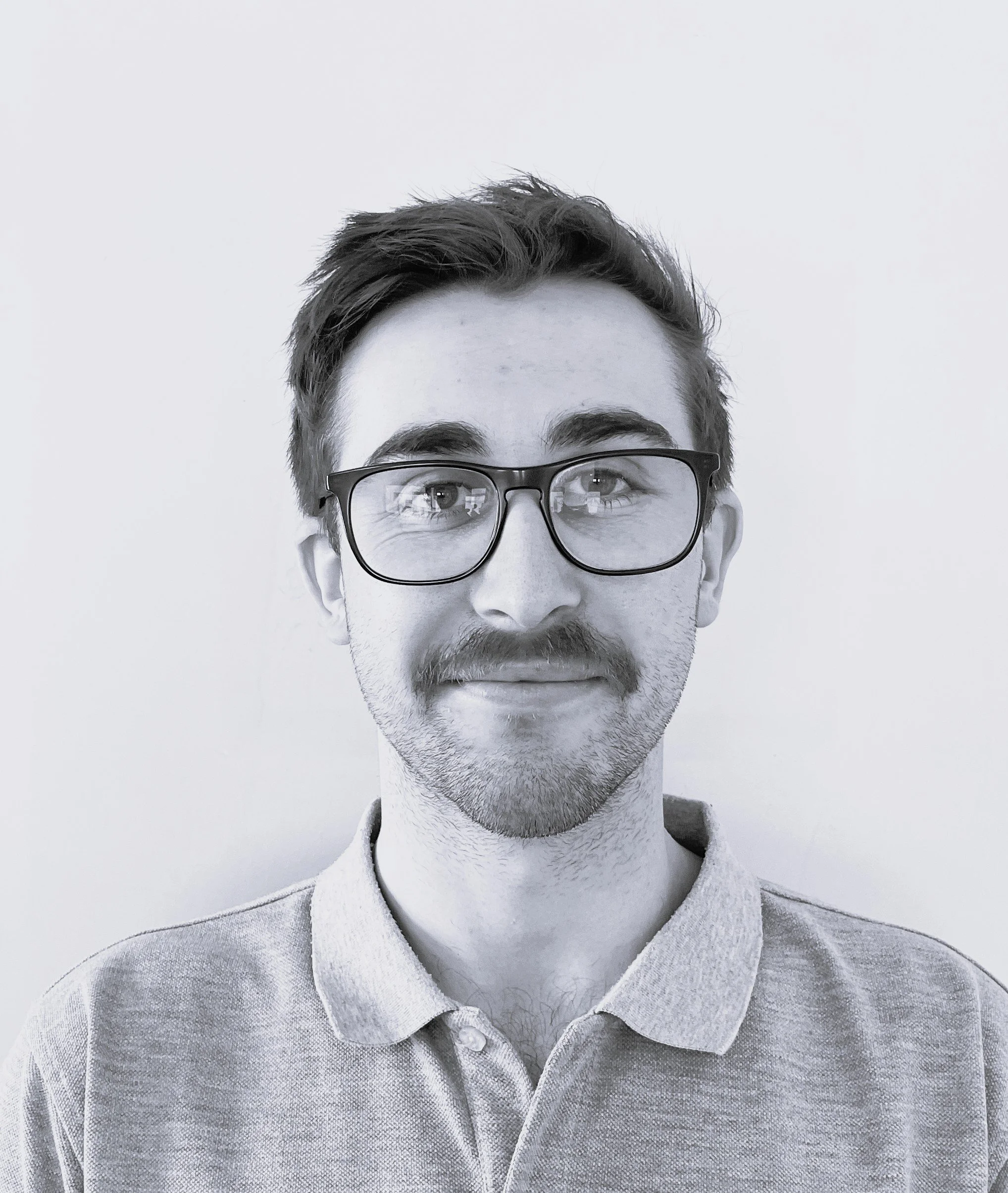 Black-and-white portrait of a young man with glasses, mustache, and beard, wearing a collared shirt, looking at the camera with a slight smile.