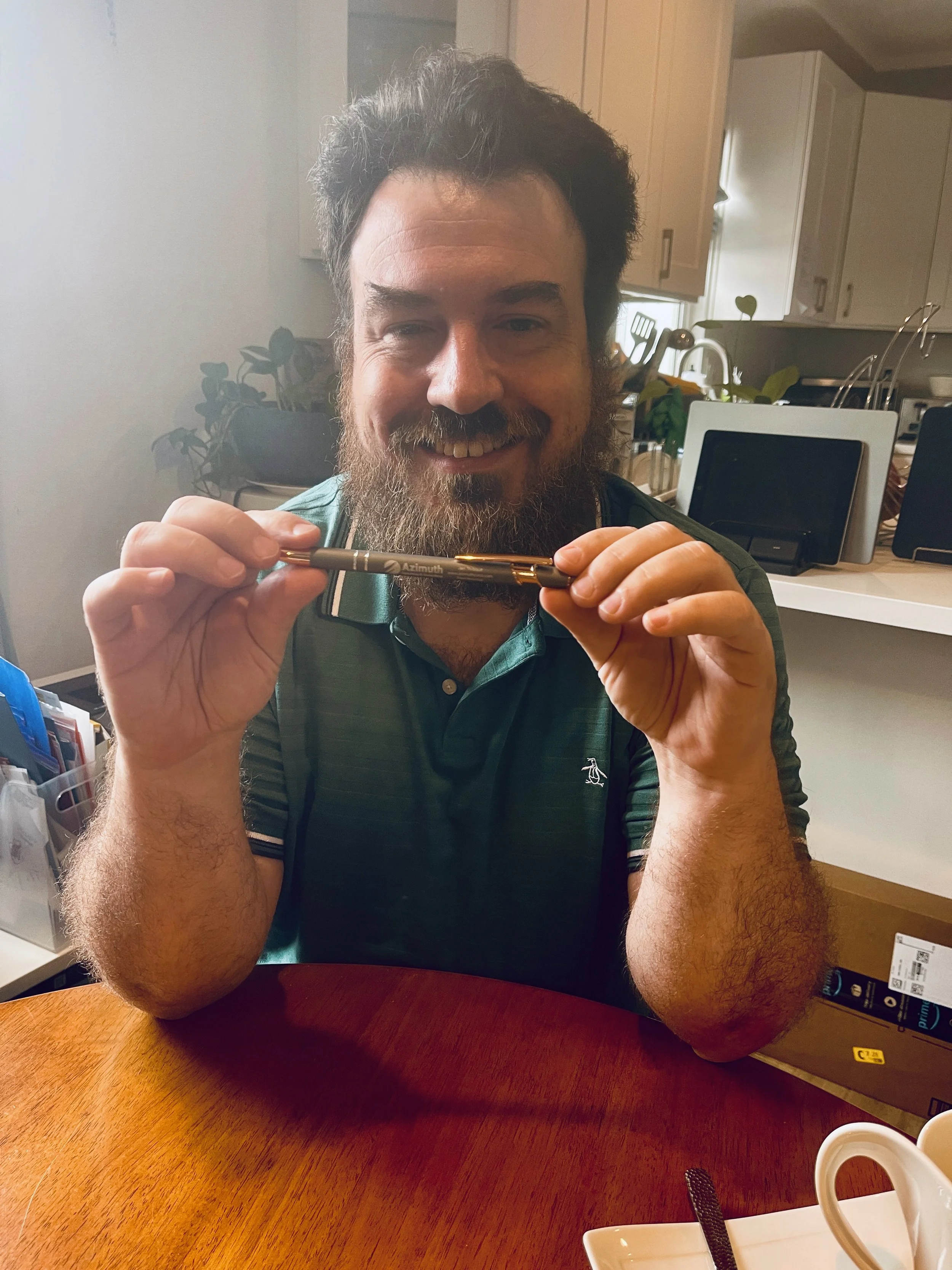A man with a beard and dark hair smiling at the camera, holding a pen horizontally in front of his face, sitting at a wooden table in a kitchen with various objects and plants in the background.