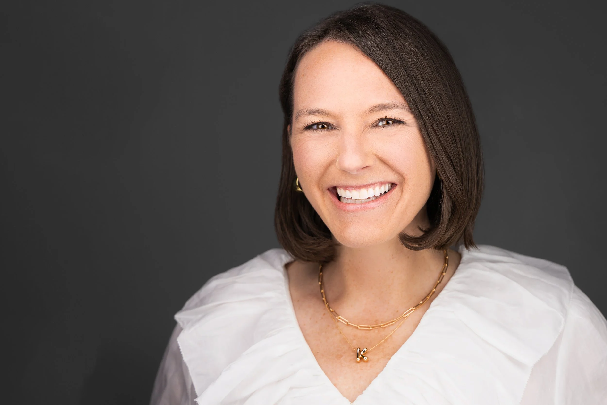 A smiling woman with shoulder-length dark brown hair, wearing a white blouse and layered necklaces, standing against a dark gray background.