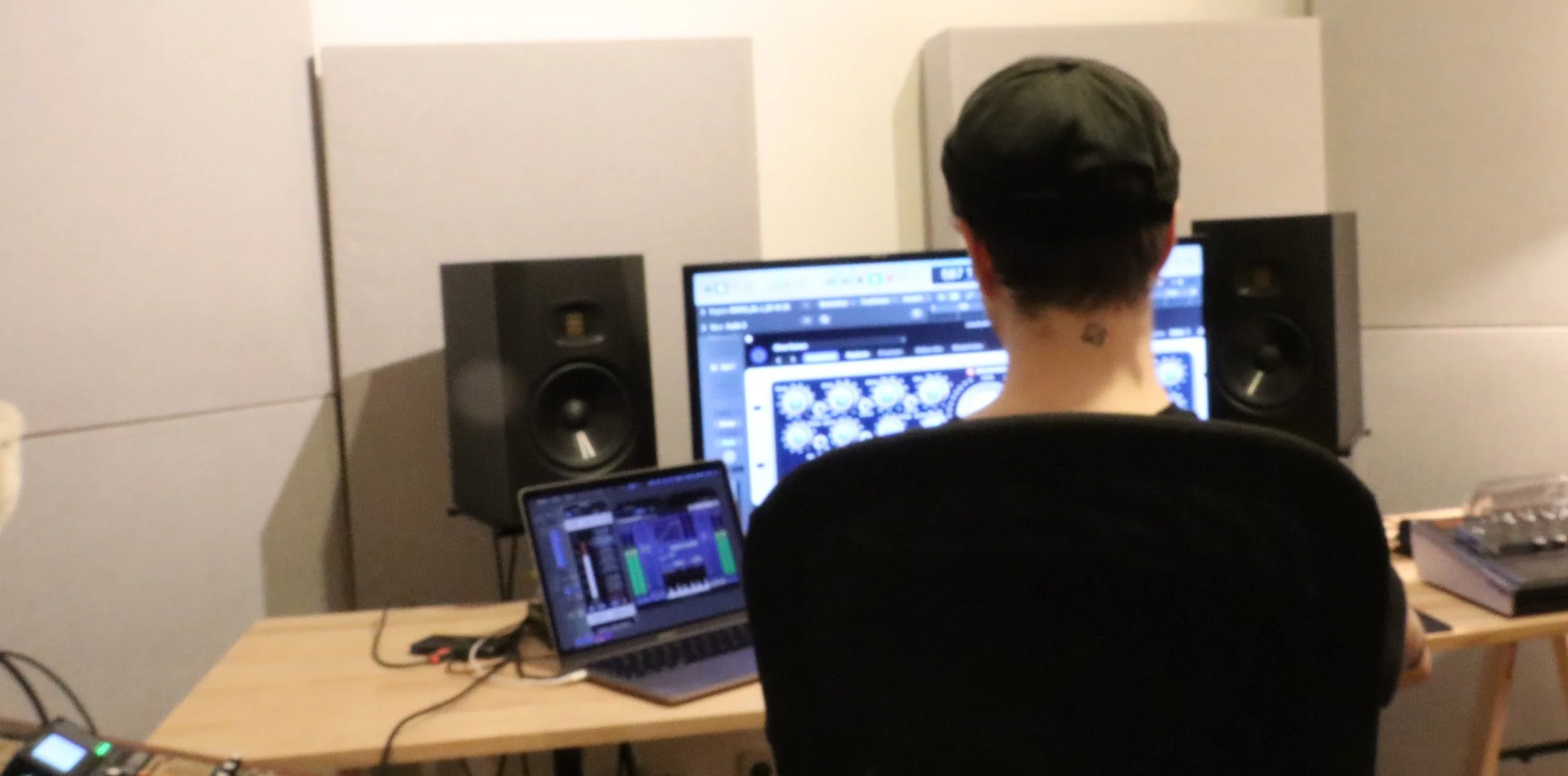 A person sitting at a music production workstation with two studio monitors, a laptop, and a large computer monitor displaying music software in a studio setting.