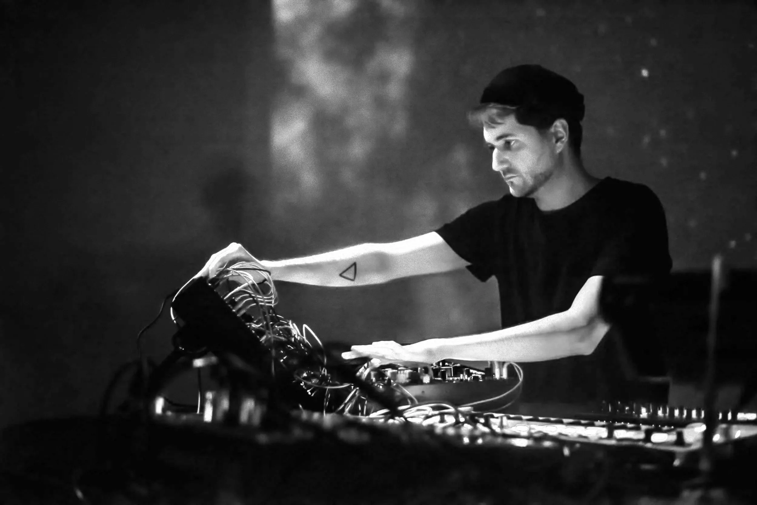 A young man in a black shirt and hat DJing with electronic equipment in a dark setting.
