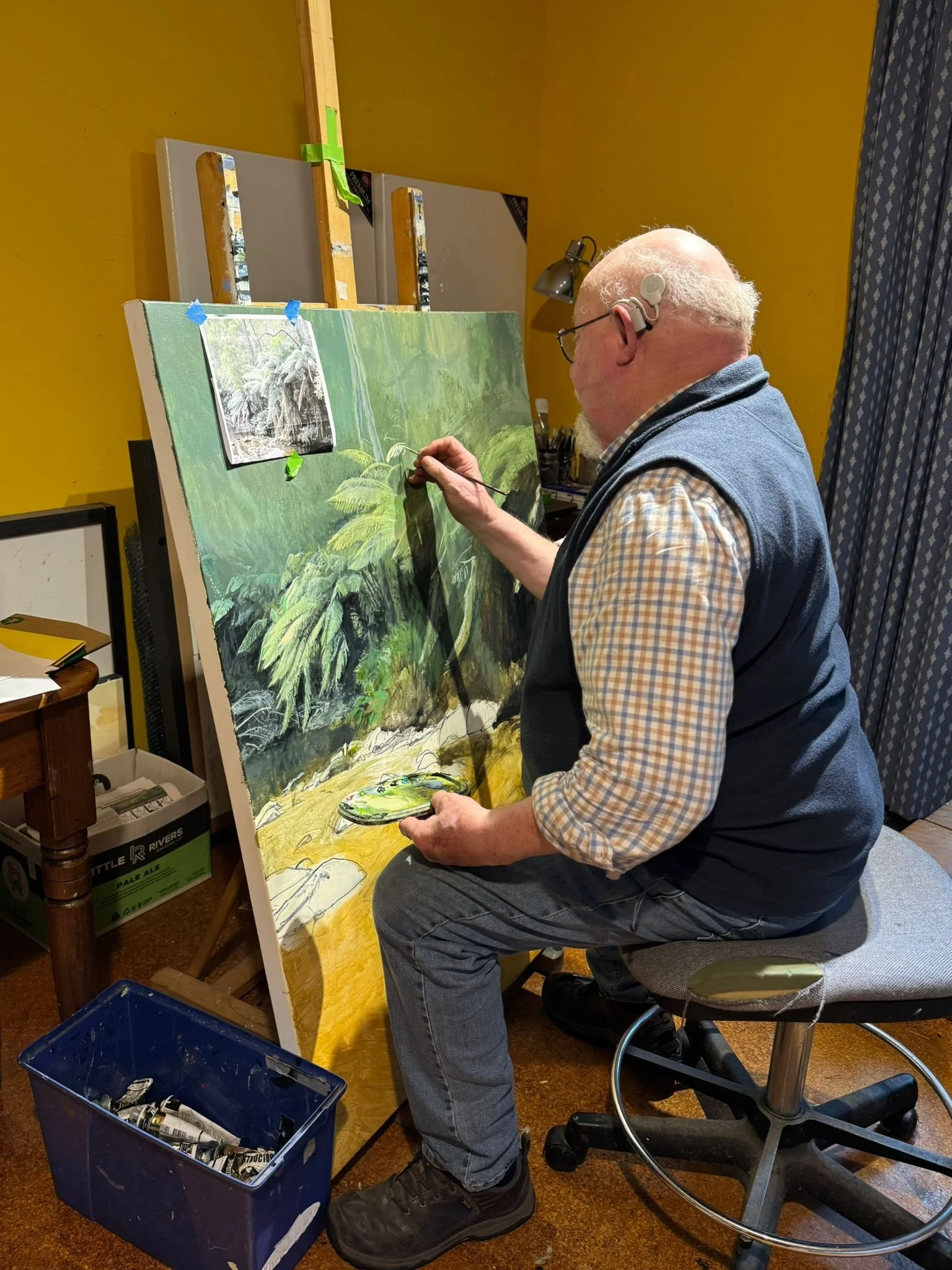 An artist painting a landscape on a large canvas in a room with yellow walls. The artist is sitting on a stool, holding a palette, and working with a brush. A reference photo is clipped near the top of the canvas.