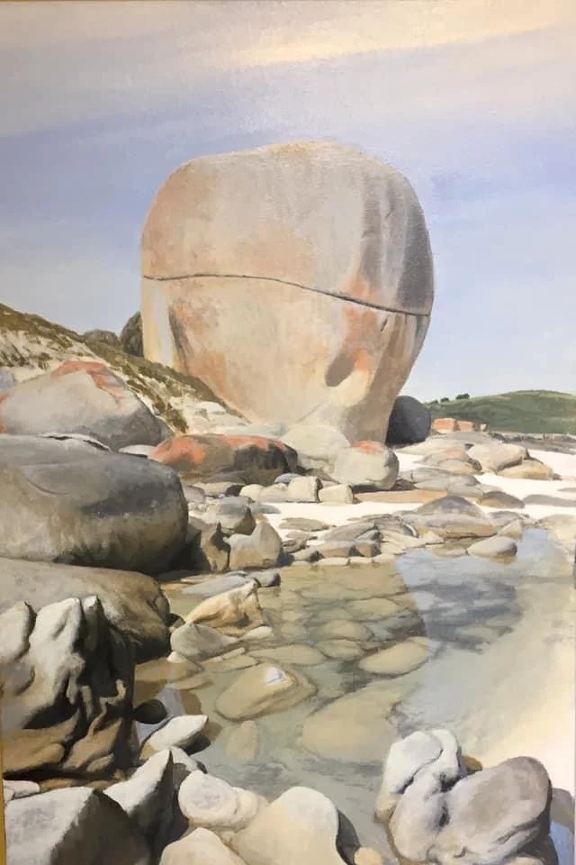 A large round boulder on a rocky riverbank with smaller stones flowing water, and a partly cloudy sky in the background.