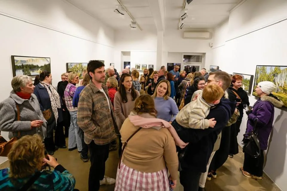 A crowded art gallery with people viewing landscape paintings displayed on white walls.