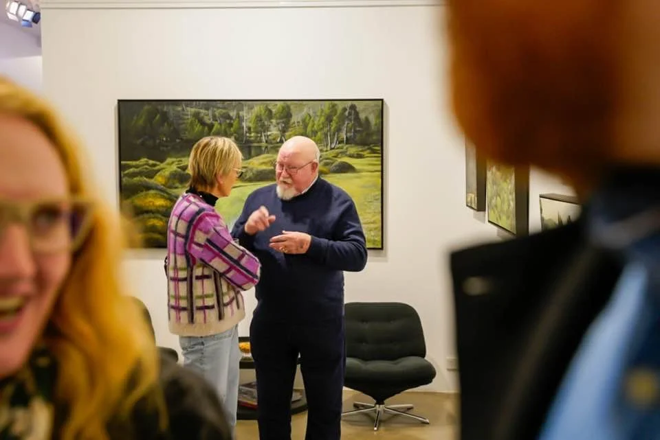 An art gallery setting with three people in the foreground and two in the background, engaged in conversation, with landscape paintings on the wall.